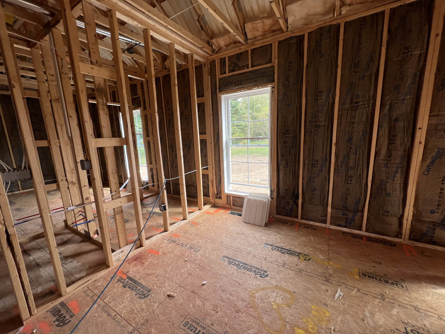 Unfinished room with exposed wood framing, window overlooking grassy field, white plastic containers on subfloor, construction materials visible