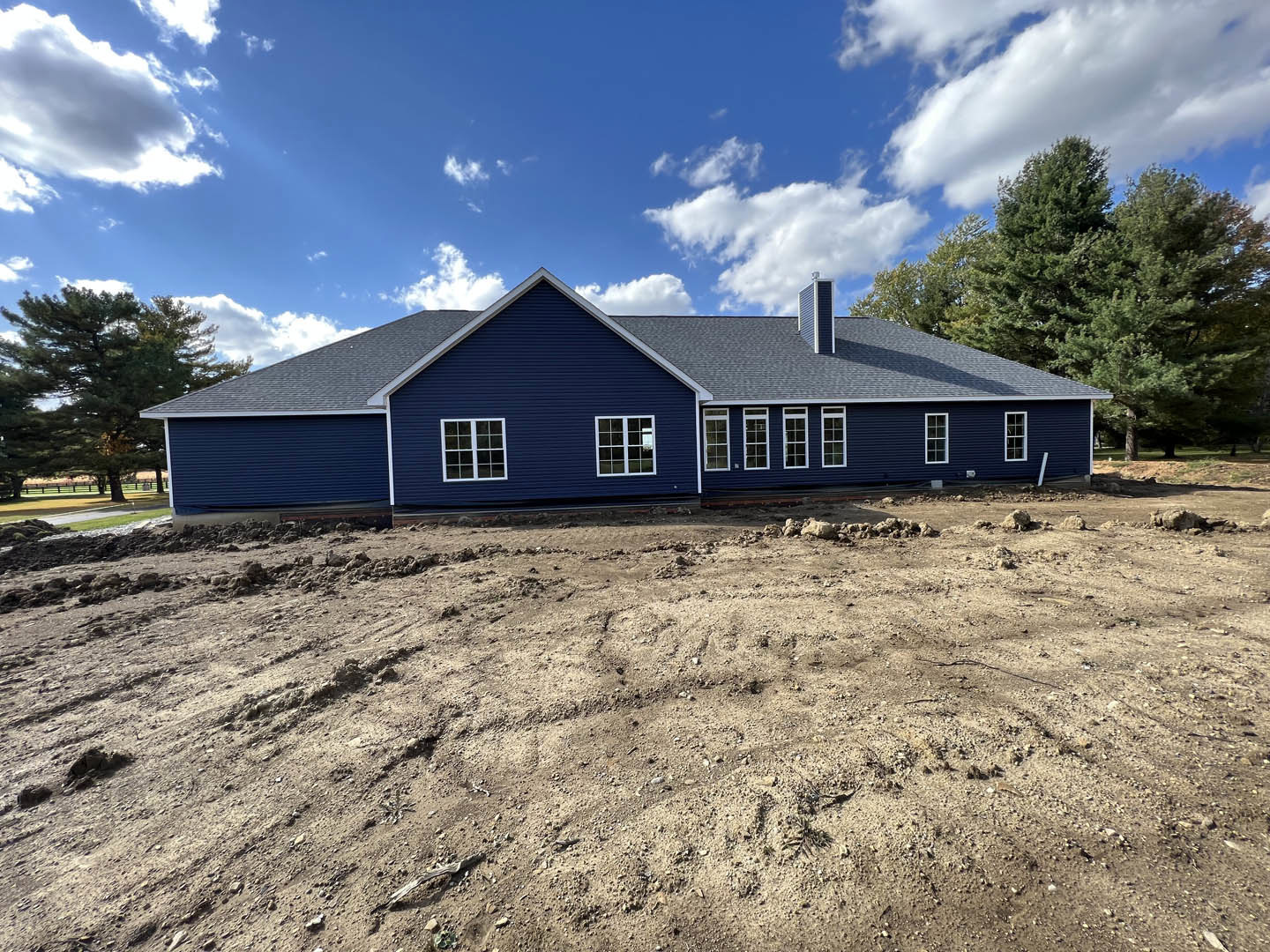 Blue house with white trim beside a dirt field, tree on the left, blue sky with scattered clouds in the background