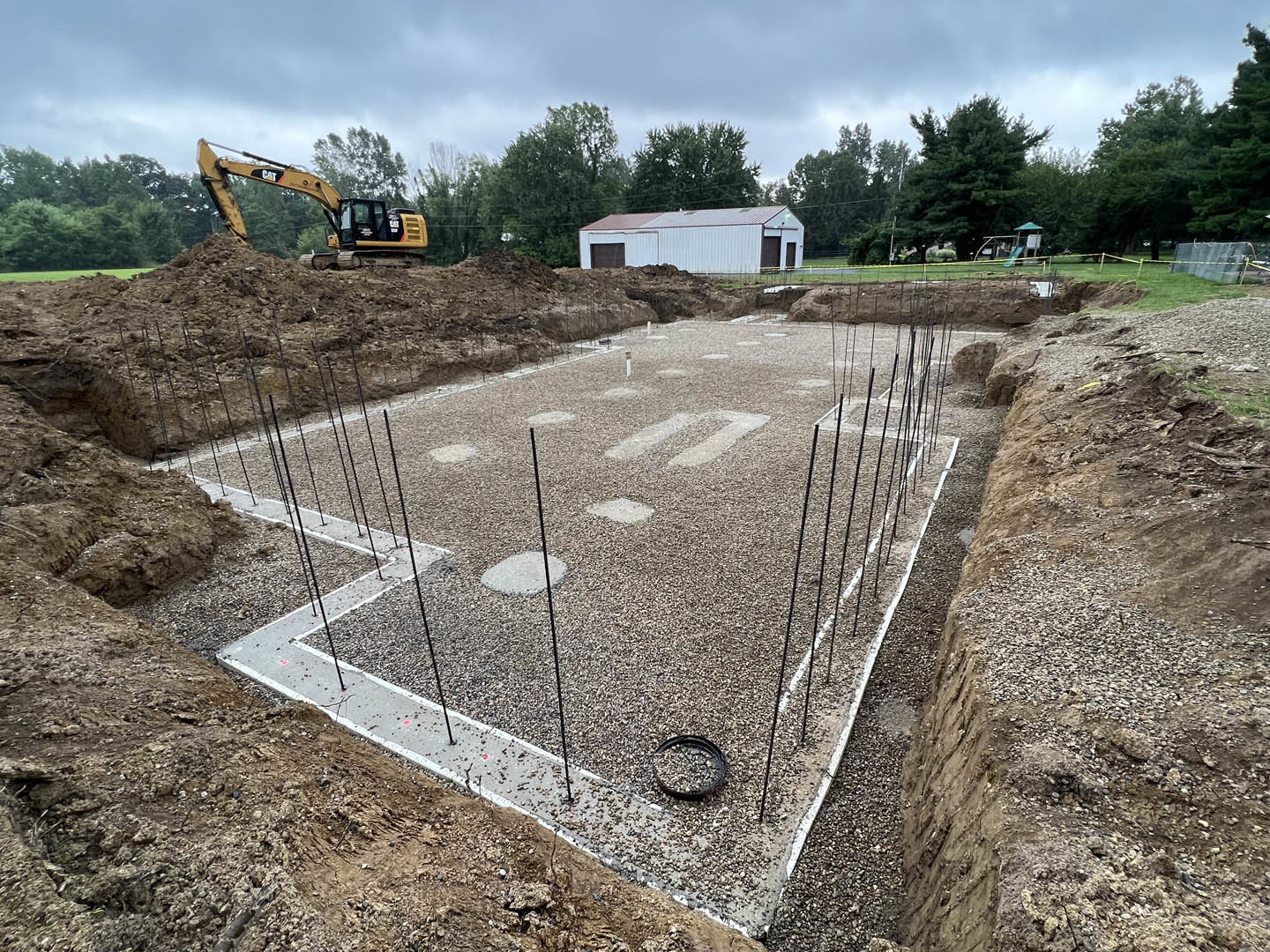 Yellow excavator digging near white building foundation, surrounded by gravel, soil, and trees on residential construction site