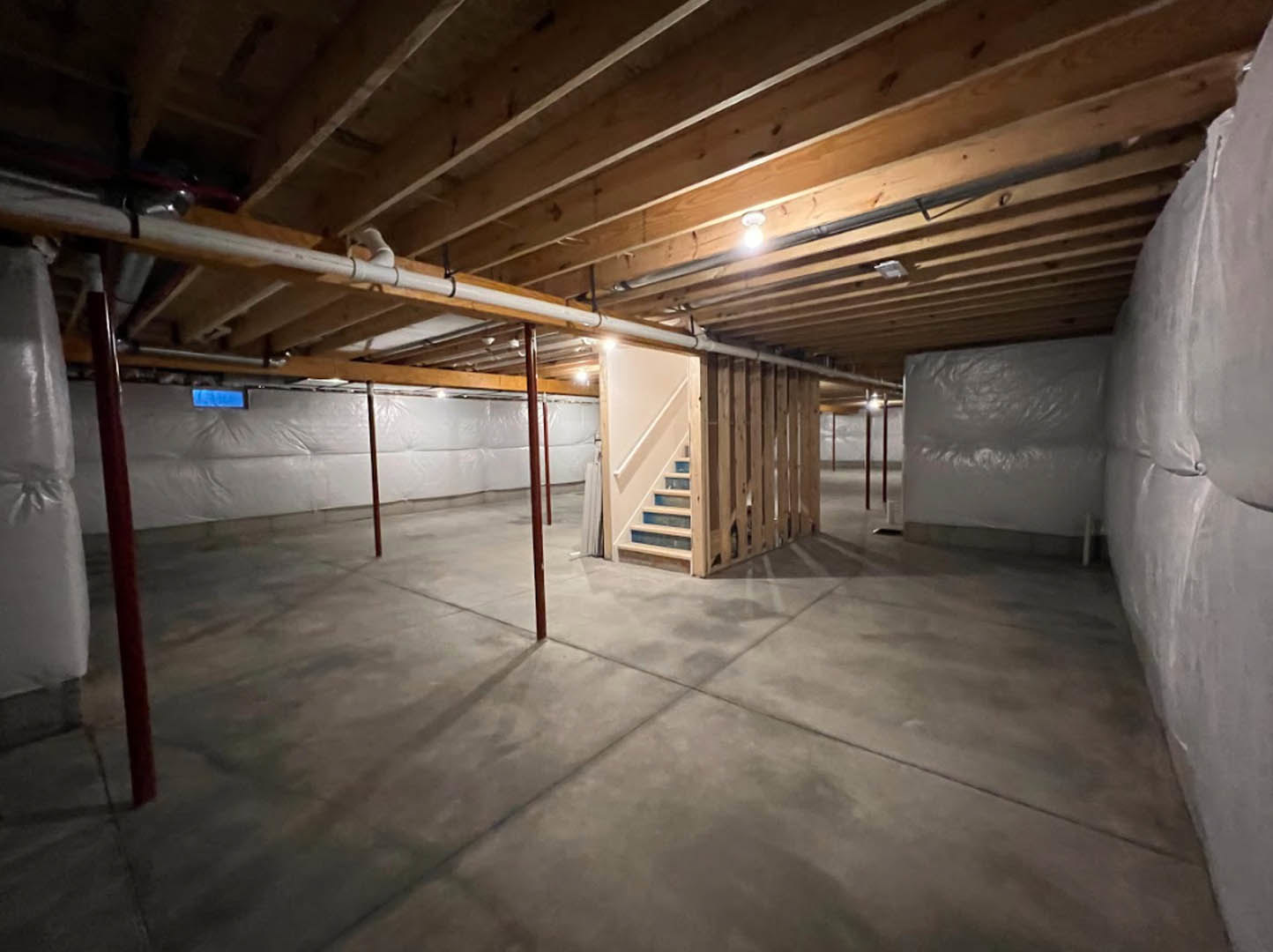 Basement with exposed wooden ceiling beams, concrete floor, staircase with wooden treads, white plastic wall wrap, visible pipes and lights overhead