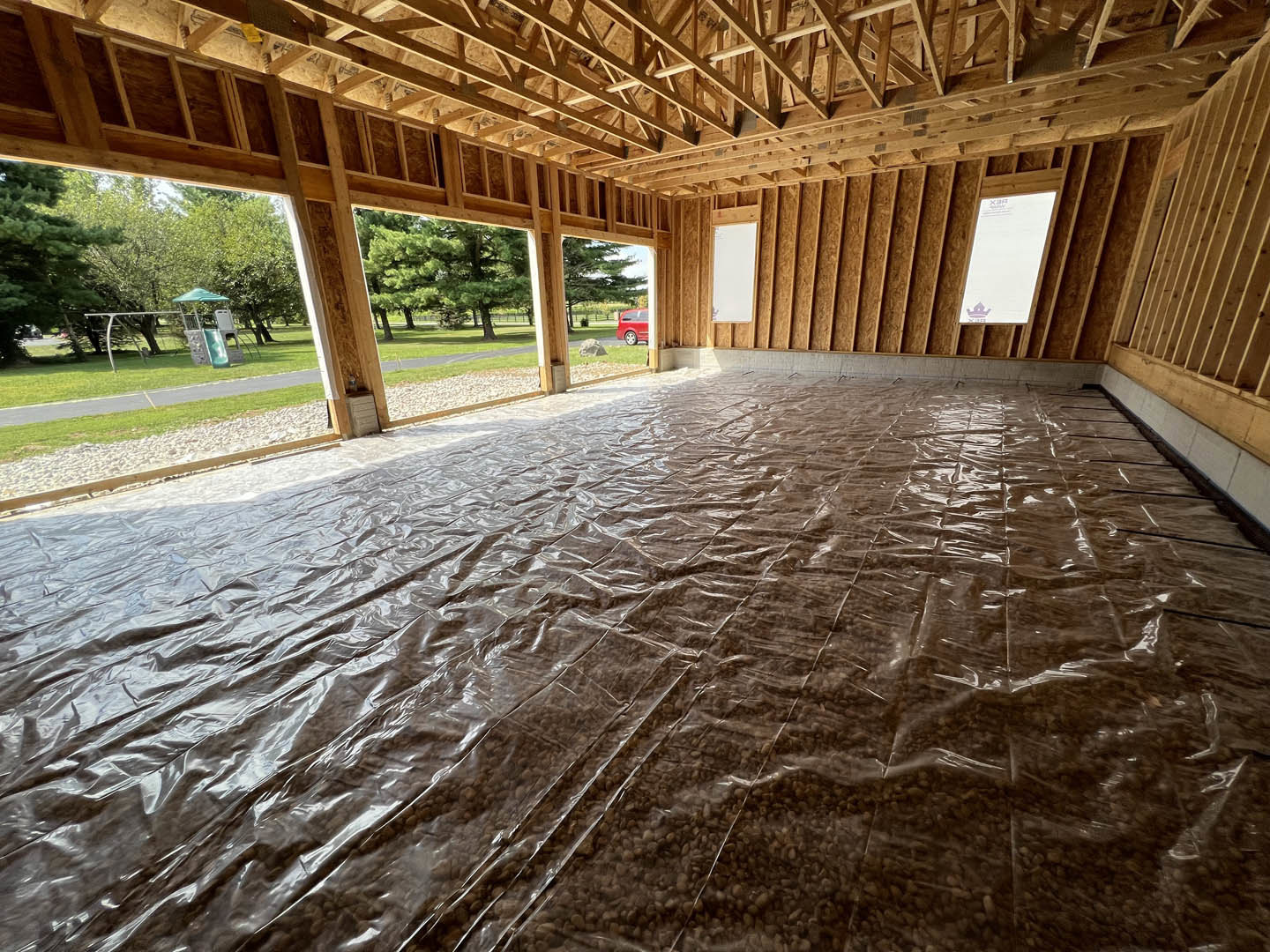 Room under construction with plastic sheeting covering the floor, exposed wooden ceiling beams, white walls with wood framing, and a rectangular object with blue text near the wall