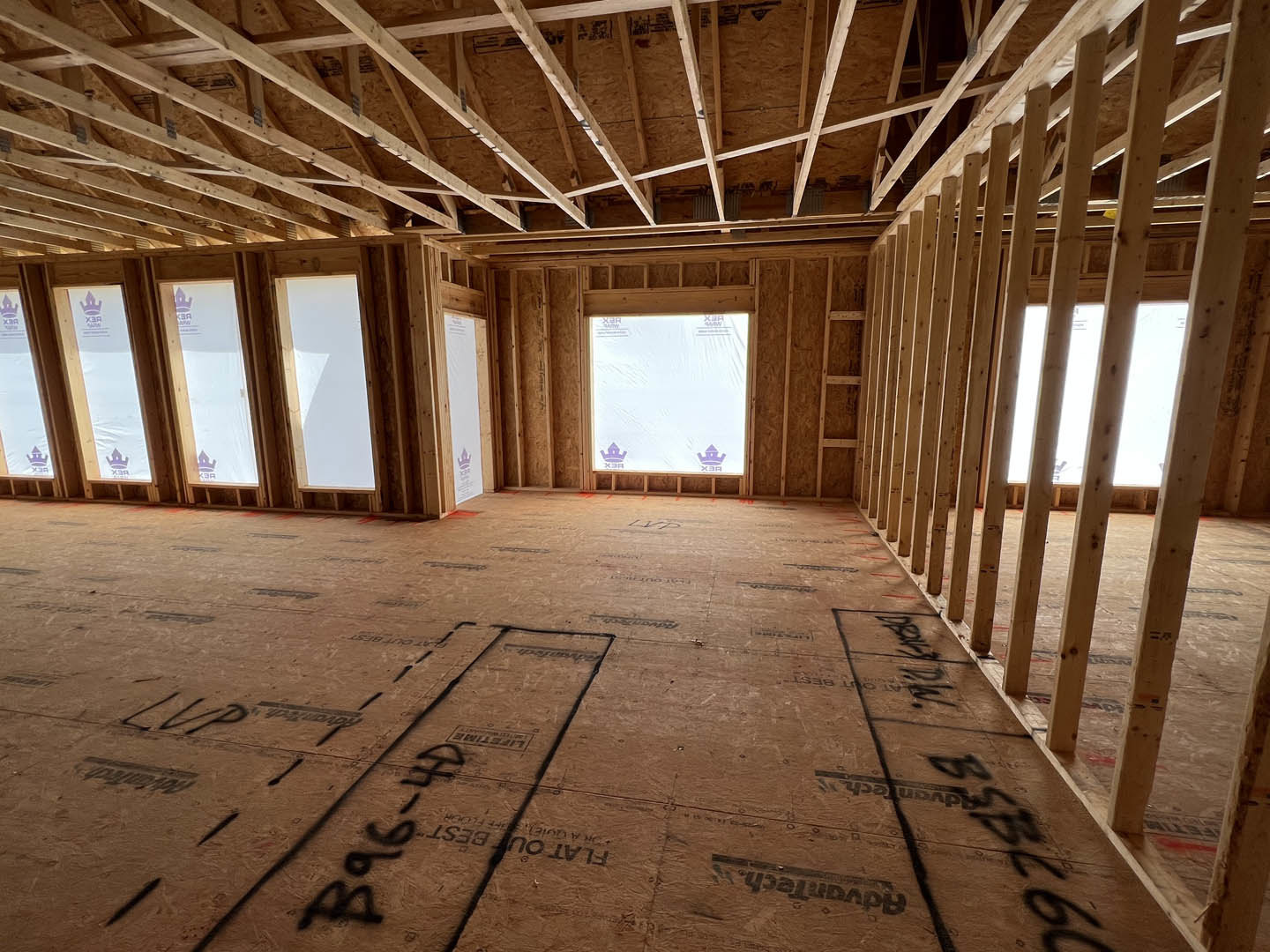 Open room with exposed wood ceiling beams, large windows, white walls, and unfinished floor marked with black writing.