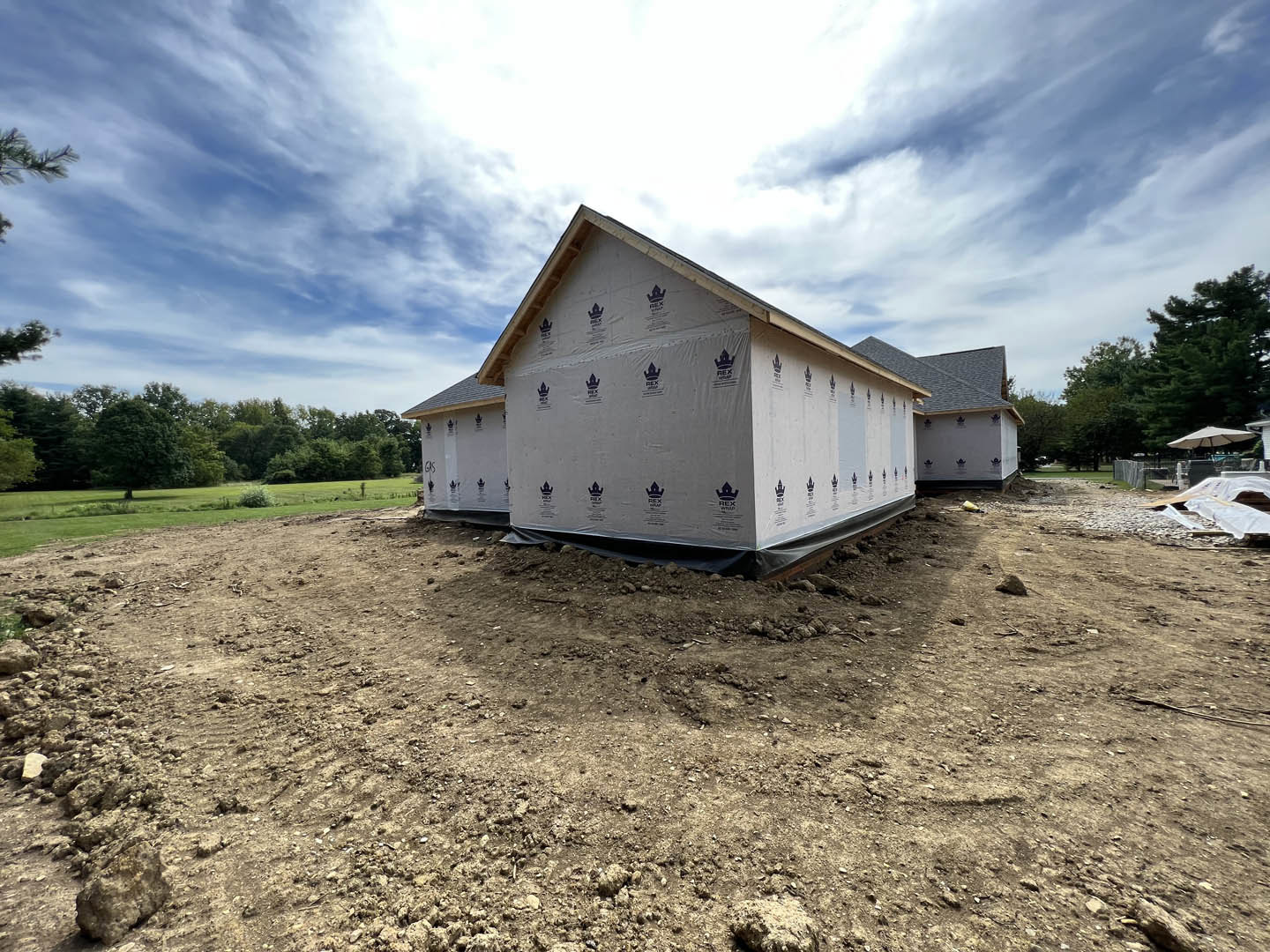 Partially built house with white sheeting covering exterior walls, grey roof wrapped in plastic, surrounded by tall trees and open ground.