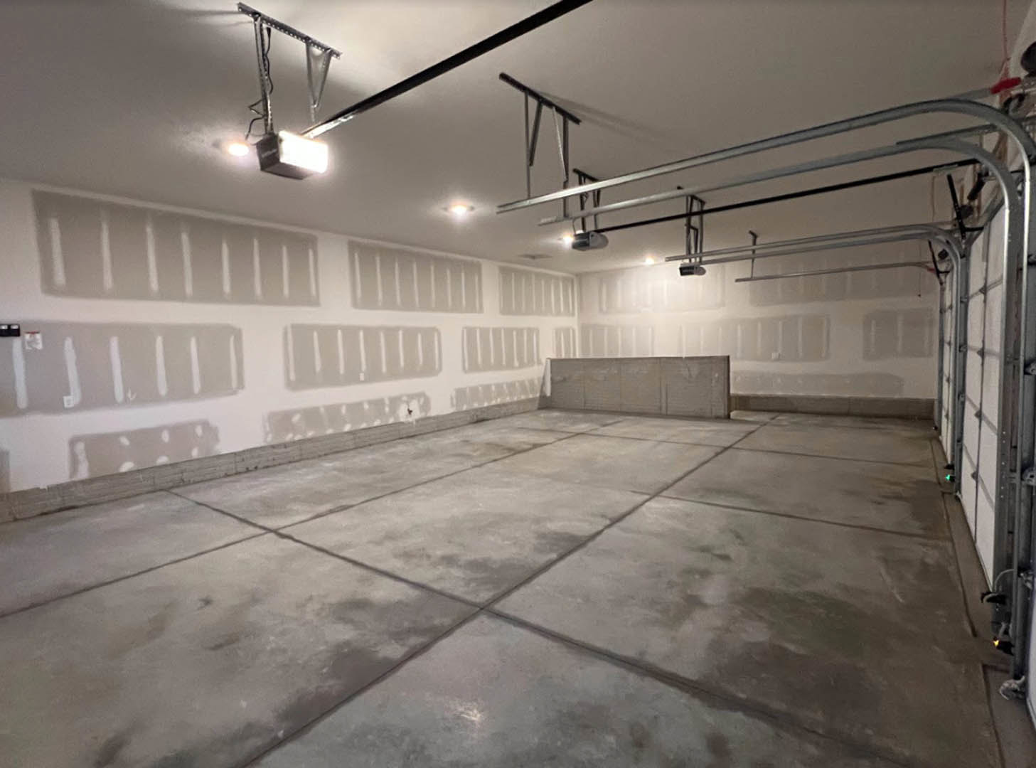 Concrete floor with white plaster walls, exposed track lighting fixtures, and visible pipe system in an empty modern interior.