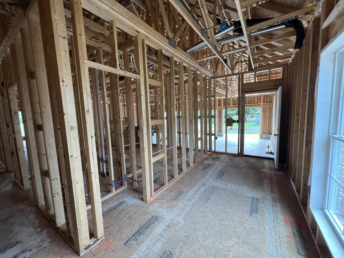Wood-framed room under construction with exposed beams, ceiling pipes, white wall with window, and doorway leading to unfinished interior