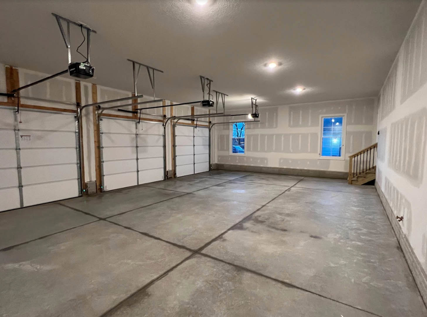 Spacious garage with grey tile flooring, white plaster walls, ceiling lights, staircase with composite material steps, window showing blue sky and tree outside