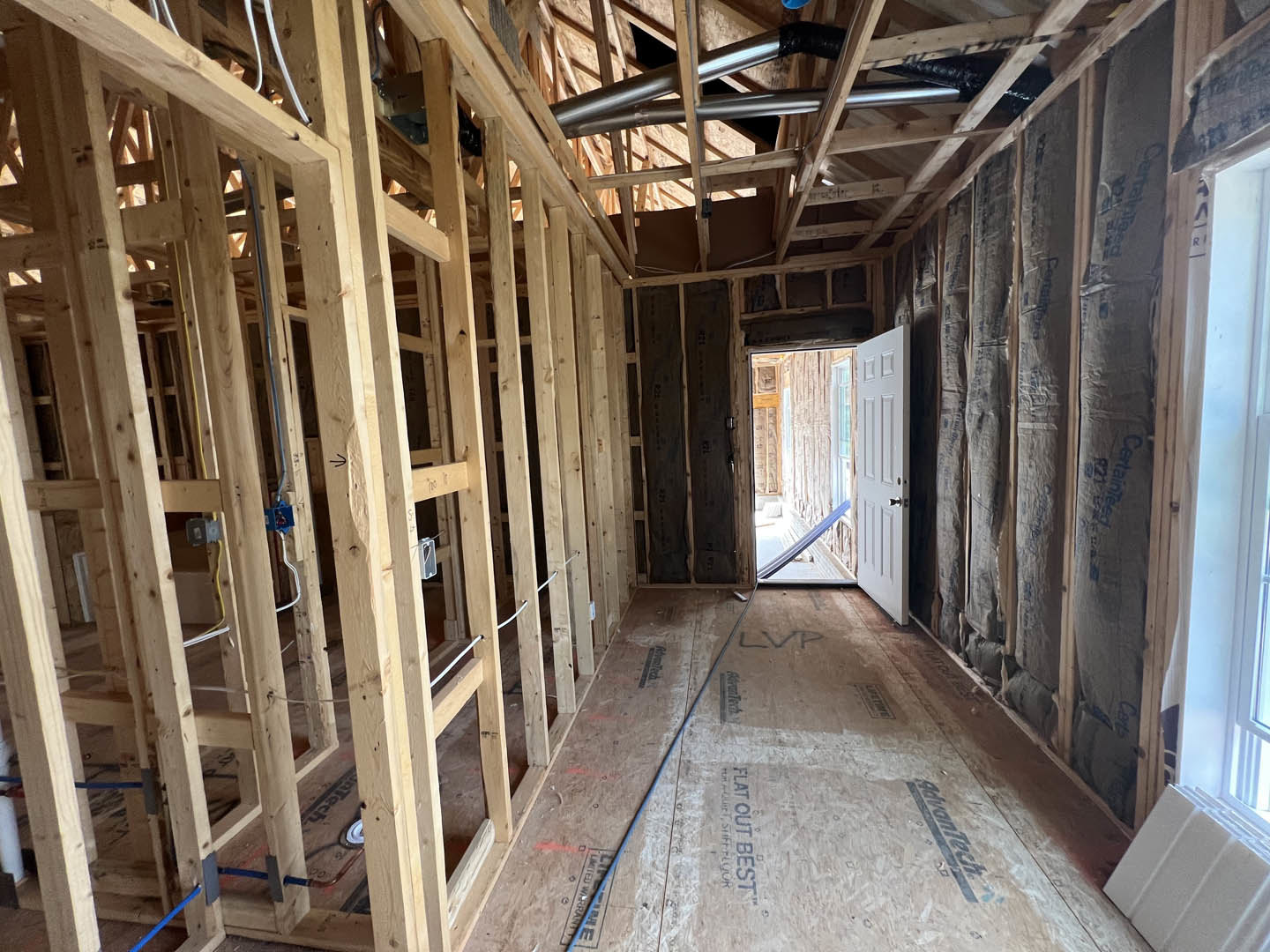 Unfinished room with exposed wood framing, white door with silver handle, hallway lined with lumber beams, metal pipes on ceiling, window, and blue chair placed centrally
