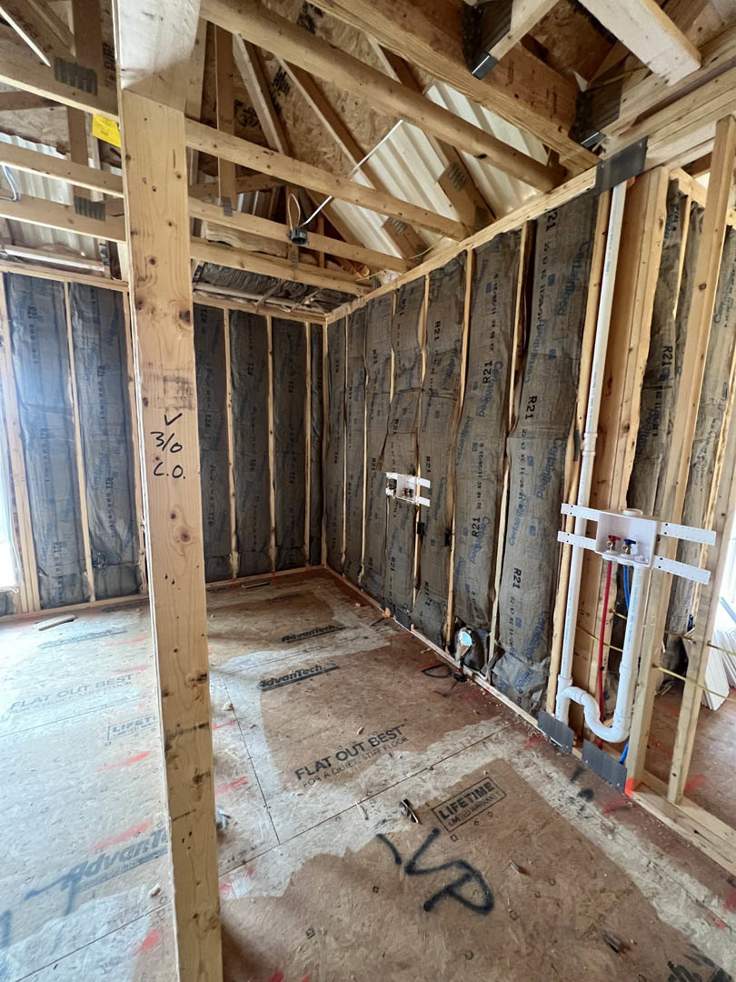 Room under construction with exposed wood ceiling beams, wall insulation, white pipes with blue and red tubing, and brown surfaces marked with black graffiti.