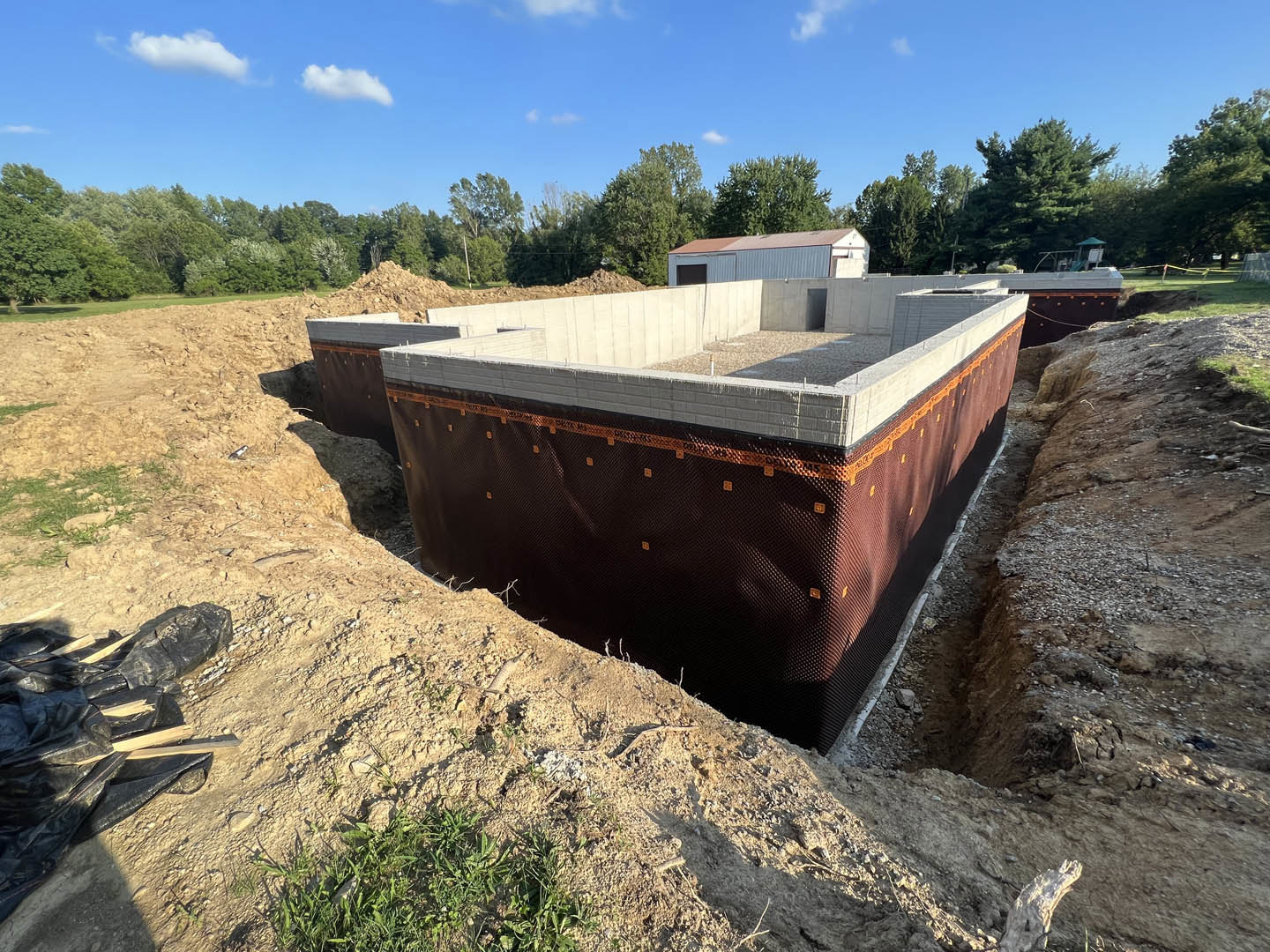 Concrete house foundation under construction with brown tarp cover, grassy foreground, blue sky with scattered clouds, trees in background