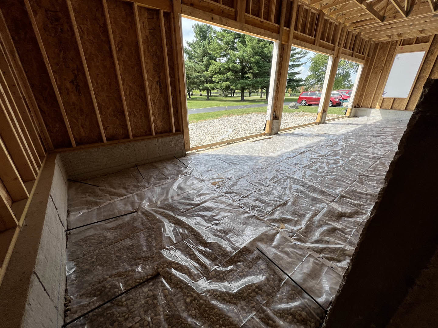 Unfinished interior with exposed wooden ceiling beams, plastic sheeting covering the floor, large windows overlooking trees, and construction materials scattered throughout