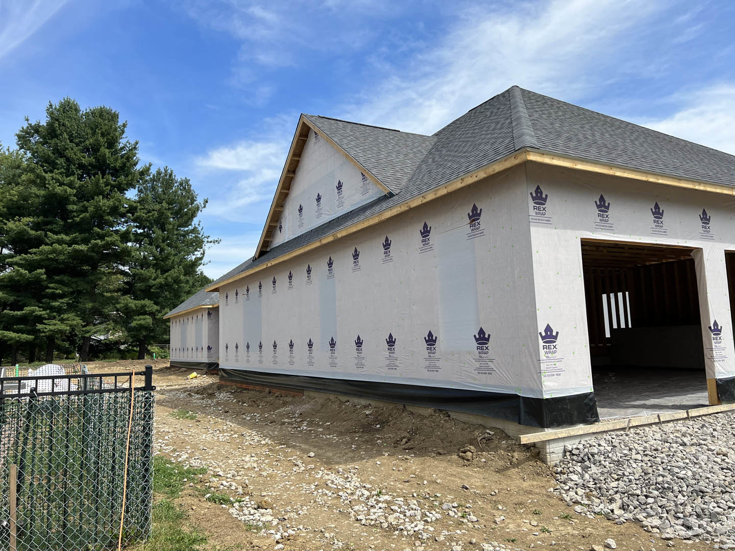 Partially built house with exposed framing, wire fence in foreground, large leafy tree, blue sky, white tent canopy, pile of rocks on gravel driveway