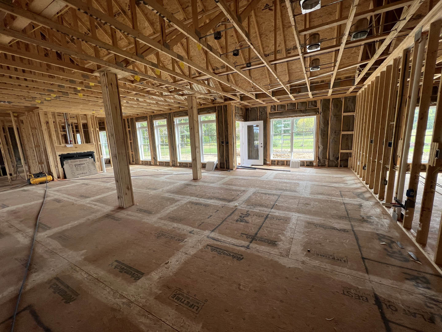 Living room with exposed wood ceiling beams, large window overlooking a grassy field, glass door, and unfinished floor marked with black construction writing.