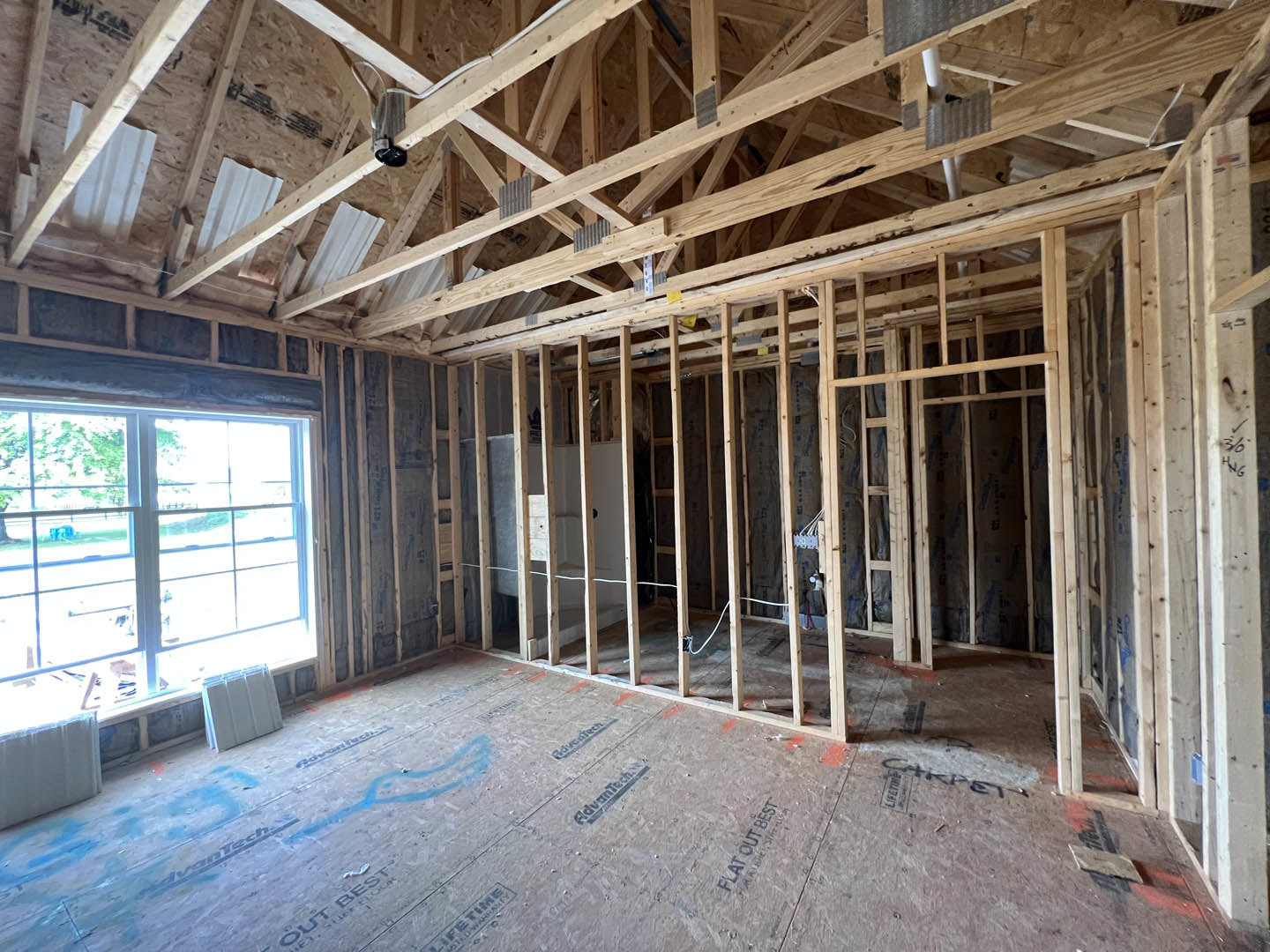 Living room with exposed wood ceiling beams, white-framed window, light-colored walls, and unfinished flooring.