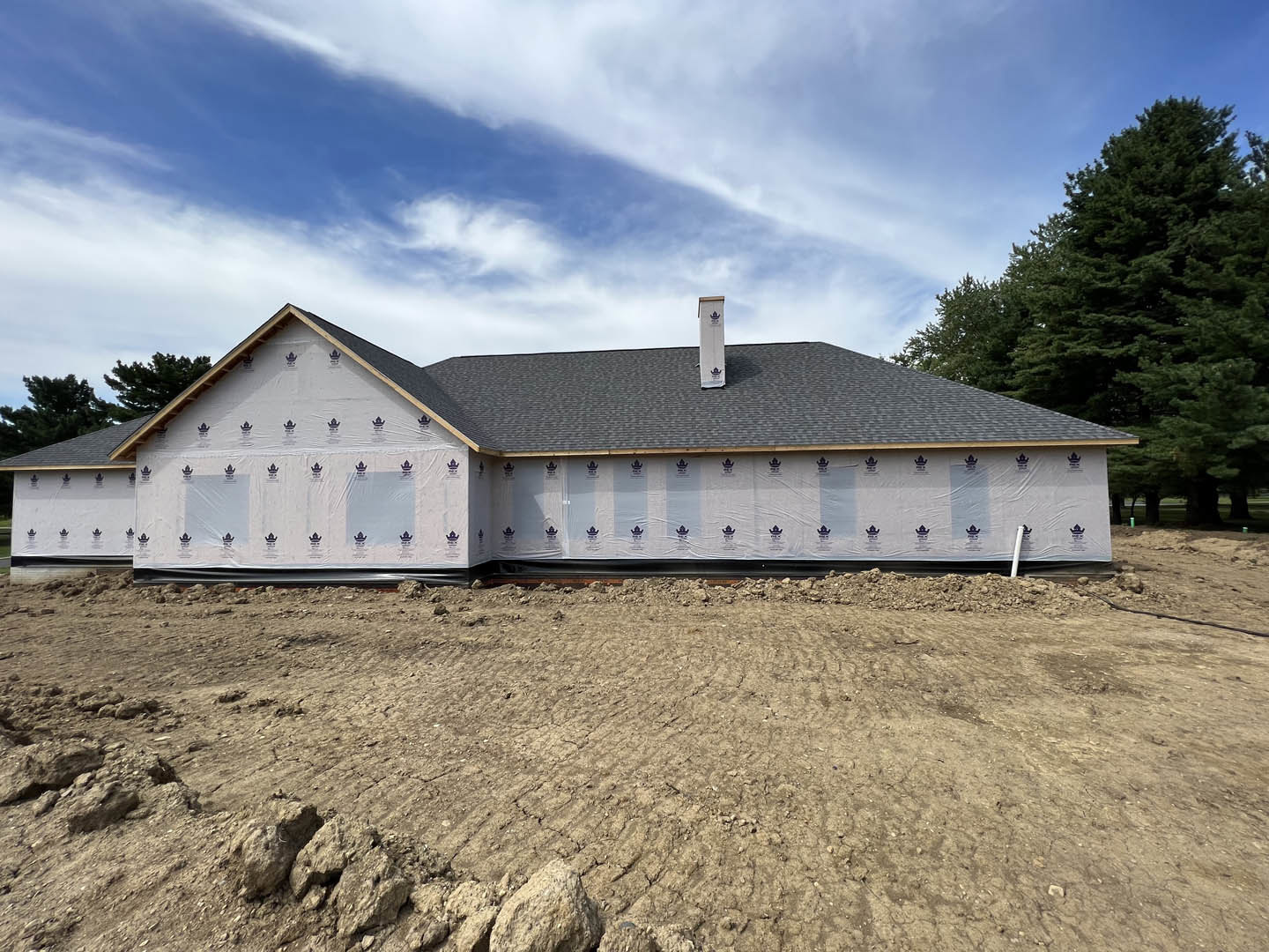 Partially built house with plastic sheeting covering exterior walls, exposed roof, chimney, dirt field with rocks, and mature trees under cloudy sky