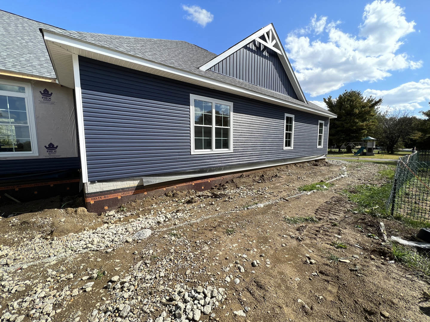 Modern home exterior with large windows, dirt path leading to entrance, unfinished landscaping with rocks and soil, blue sky overhead, partial view of white fence and construction