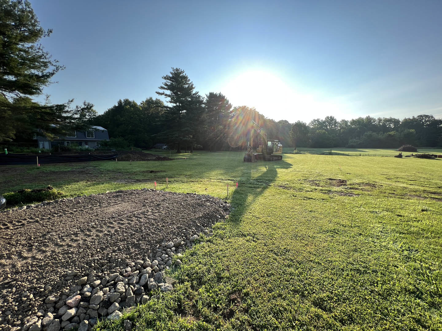 Bulldozer parked on dirt and rocky construction site, surrounded by grassy field and clusters of leafy trees, sunlight filtering through branches, house partially visible in