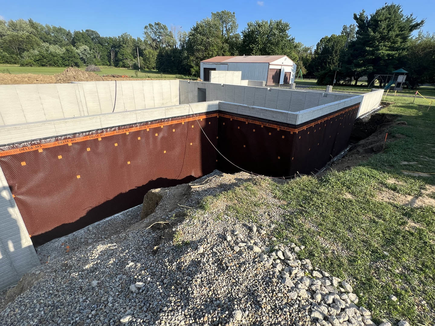 Partially built house with blue and brown roof, pile of gravel in foreground, grassy yard, trees and playground set in background