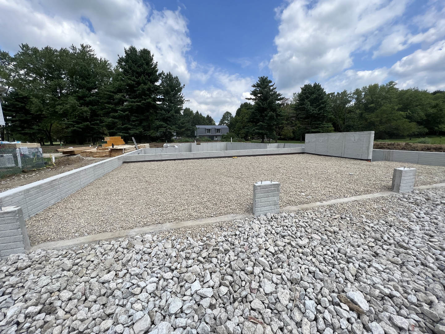 Partially built house with exposed concrete block walls, metal rebar, gravel and rocks piled beside the foundation, curb, and scattered trees under a blue sky with clouds