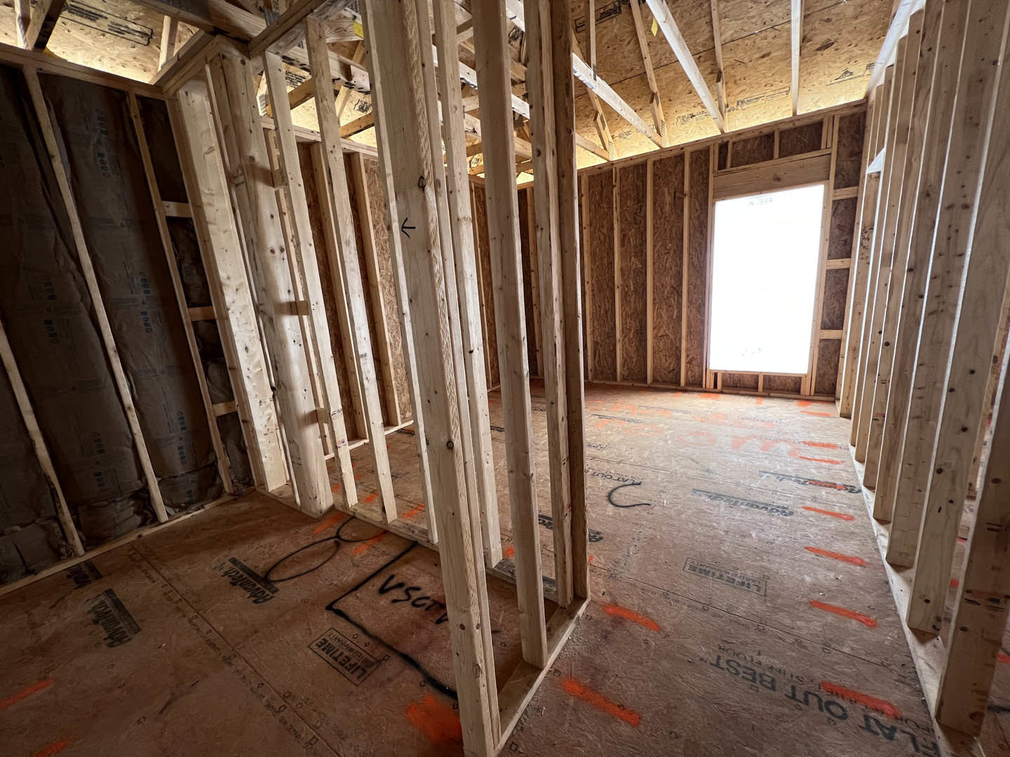 Exposed wood framing and beams in a partially constructed room with building insulation and composite materials stacked on the floor, illuminated by overhead white light.