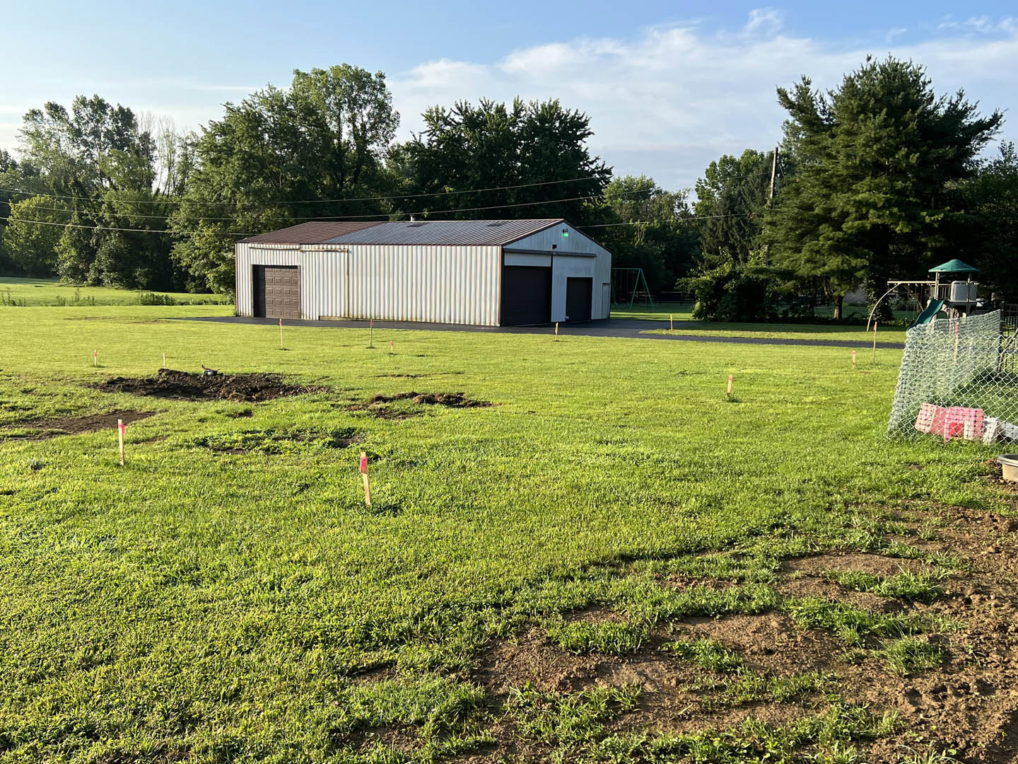 Modern farmhouse with white siding and dark roof set in a grassy field, surrounded by trees and a chain link fence, brown garage door visible on the side.