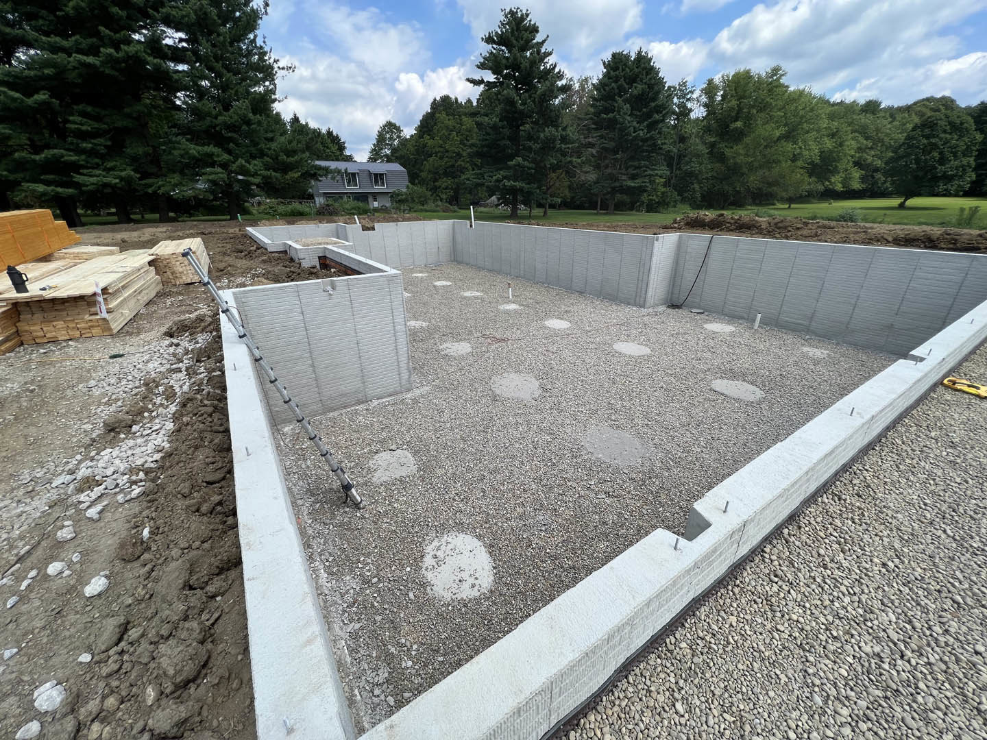 Concrete foundation surrounded by gravel, stack of wood planks, tall leafy tree, house with roof and walls in the background, cloudy sky overhead