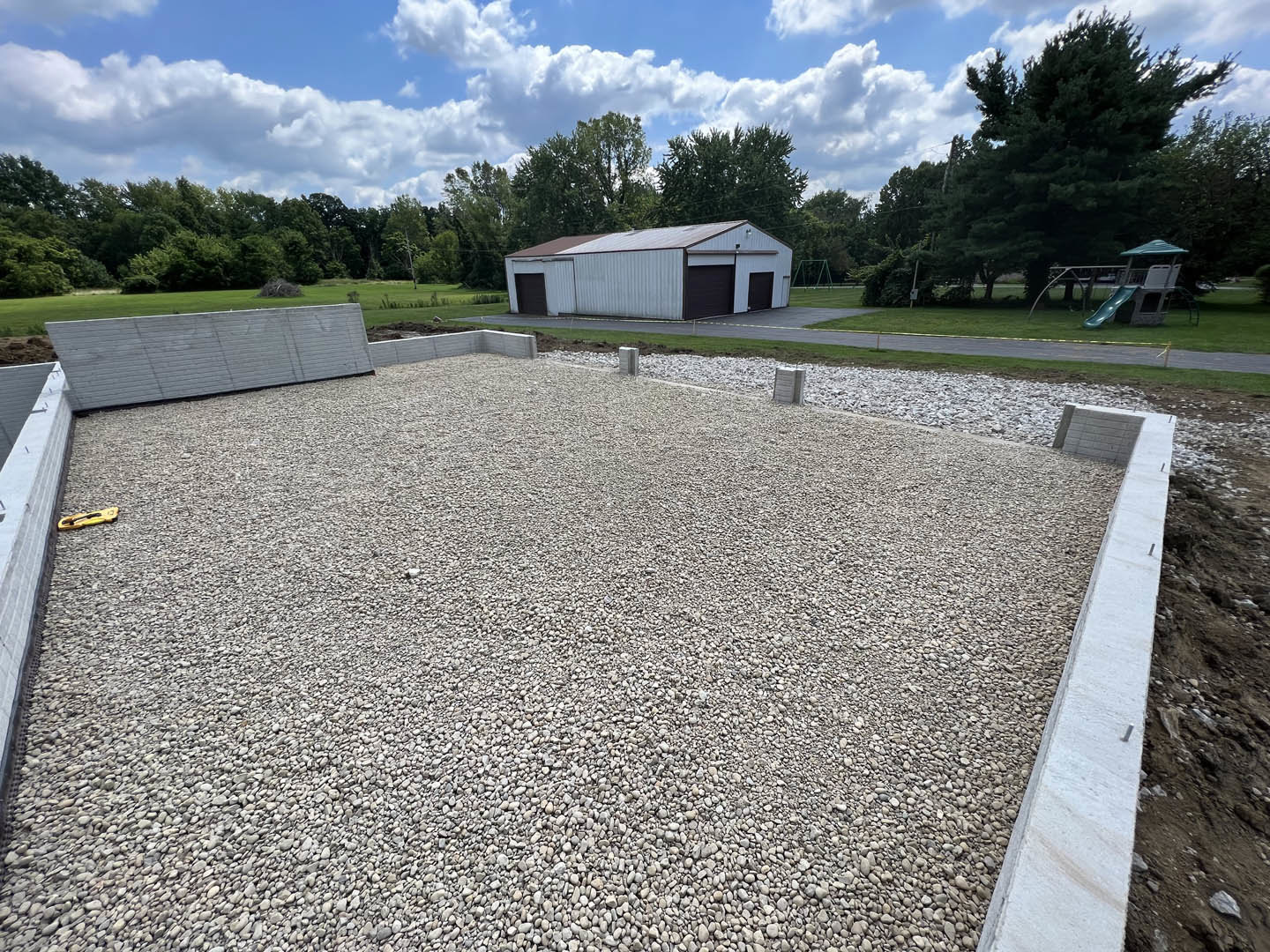 White and brown house with attached garage, gravel driveway, green lawn, trees, and blue sky with clouds