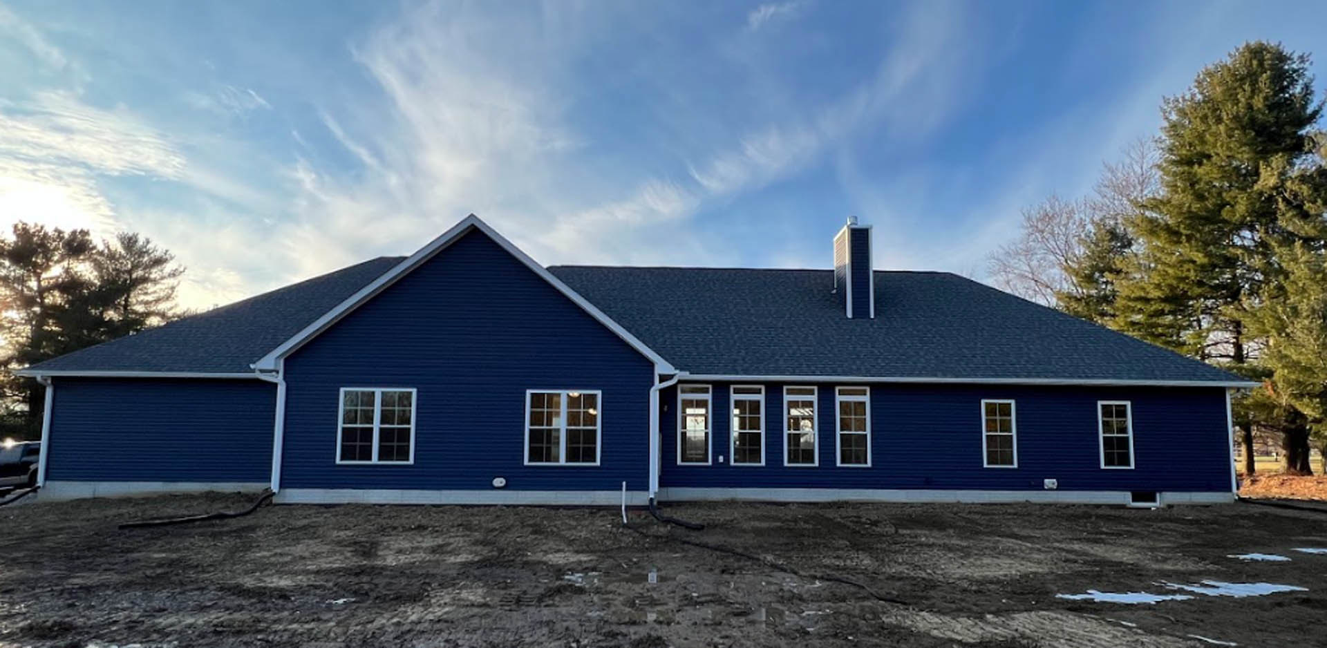 Blue house with matching blue roof, white trim around windows and doors, large window with white frame, tree in background, cloudy sky above, landscaped ground in foreground