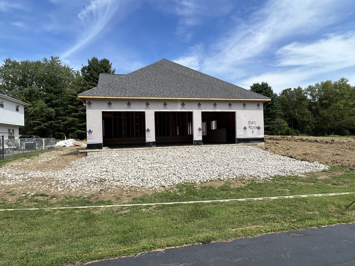 Partially built house with exposed roof trusses, gravel driveway, grassy yard marked by yellow tape, white curtained window, surrounded by trees under cloudy sky