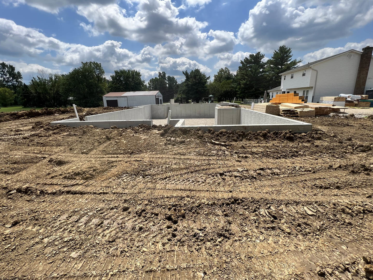 Large concrete foundation surrounded by dirt and construction materials, wood pallets stacked near a partially built wall, white metal building with door in background, cloudy sky
