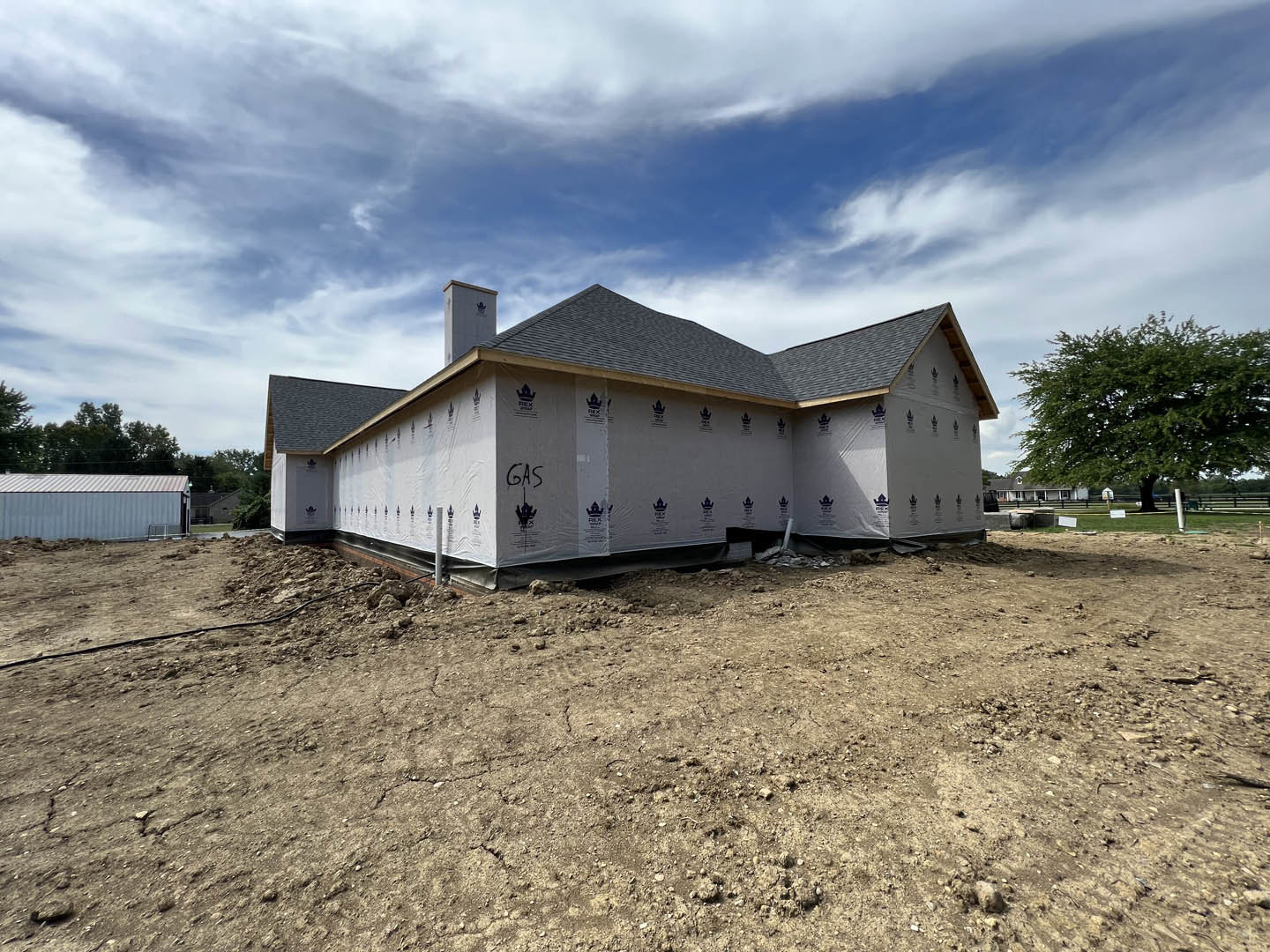 Partially built house with exposed framing, surrounded by dirt and mature trees, white construction tarp covering part of the structure, metal container with sloped roof nearby