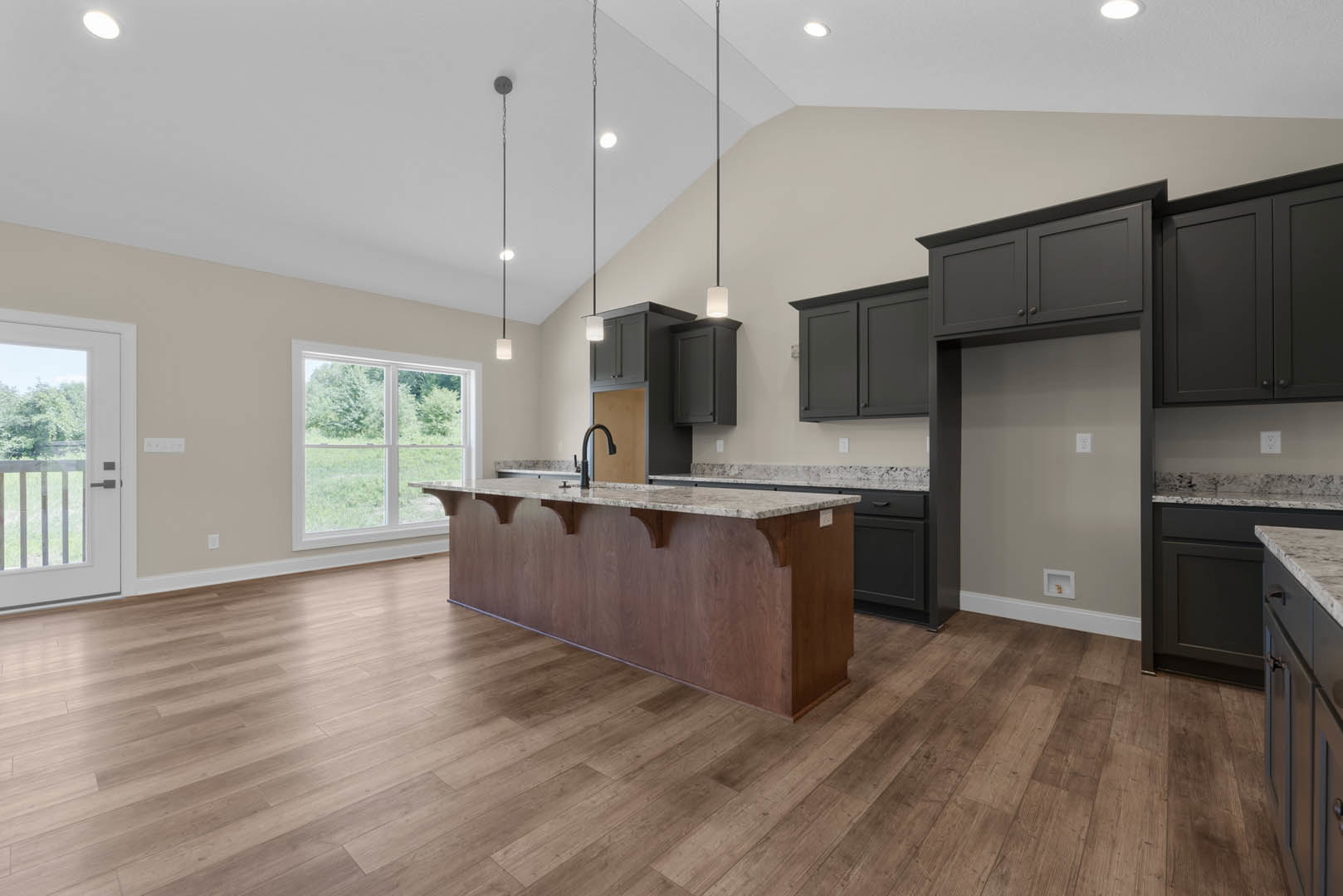 Spacious kitchen featuring a large marble-topped island, wood flooring, white cabinetry, black faucet, and a window overlooking a grassy yard.