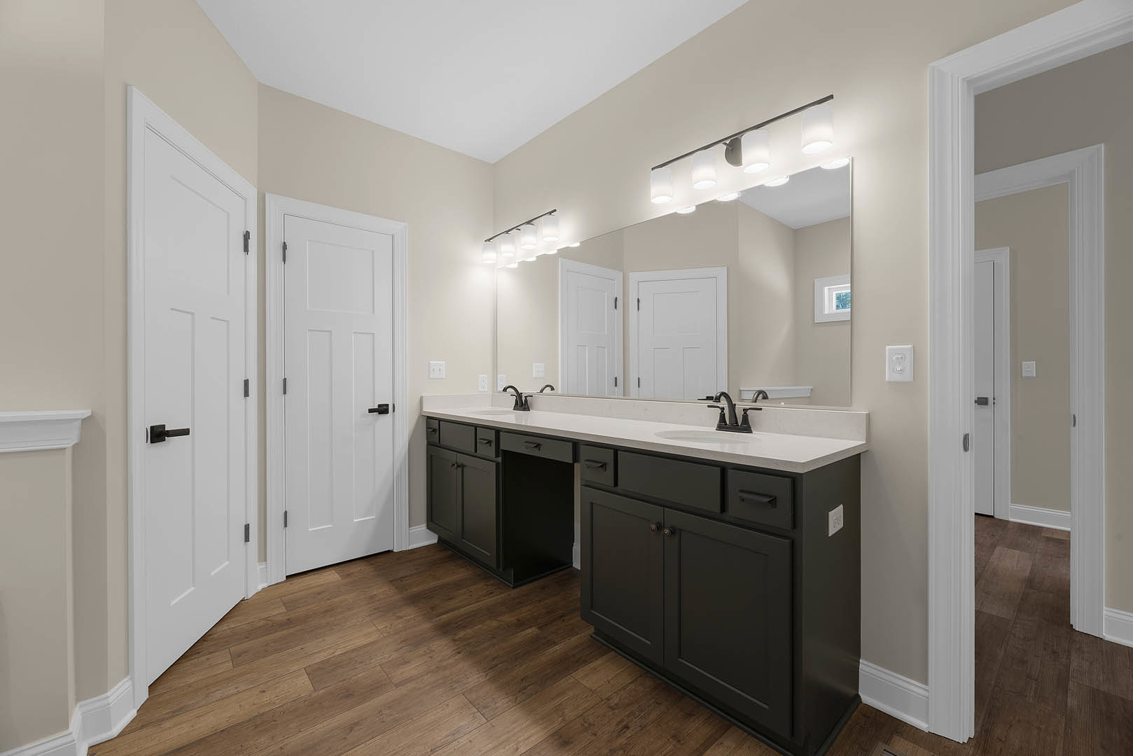 Bathroom with double white sinks set in a black vanity, wide rectangular mirror above, row of wall-mounted lights, wood flooring, white door with black handle and square frame.