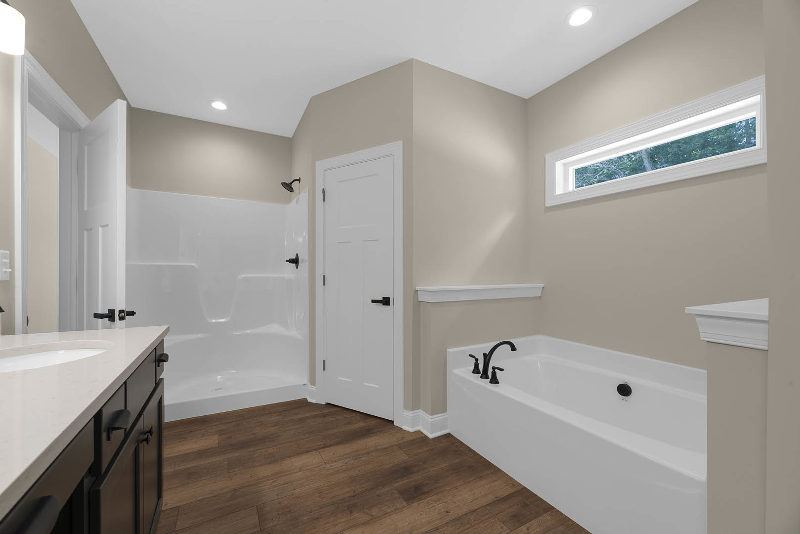 Bathroom with freestanding white bathtub and black faucets, white vanity with undermount sink, black hardware, light gray tile floor, white door, large window overlooking trees