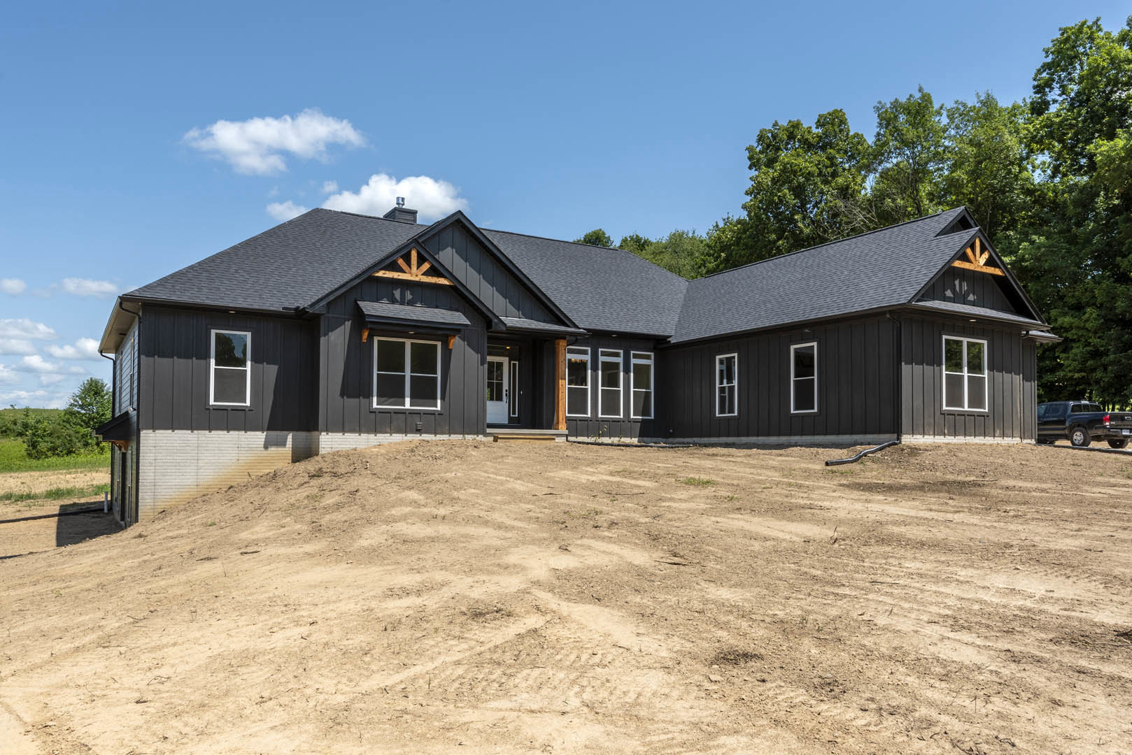 Two-story house with white trim and large windows, situated behind a dirt hill and field, black truck parked on paved road, cloudy sky overhead, trees bordering property