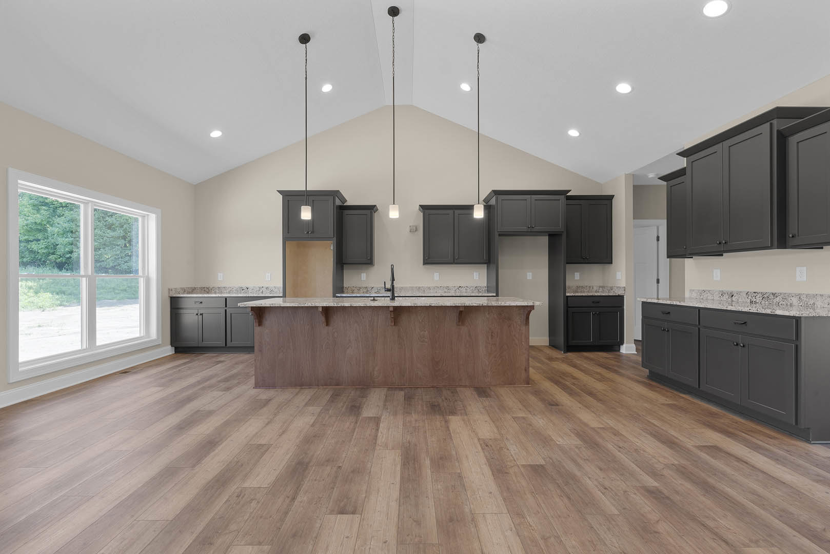 Spacious kitchen featuring a large central island with light stone countertop, wood flooring, wood-paneled accent wall, white cabinetry, stainless steel appliances, and a window