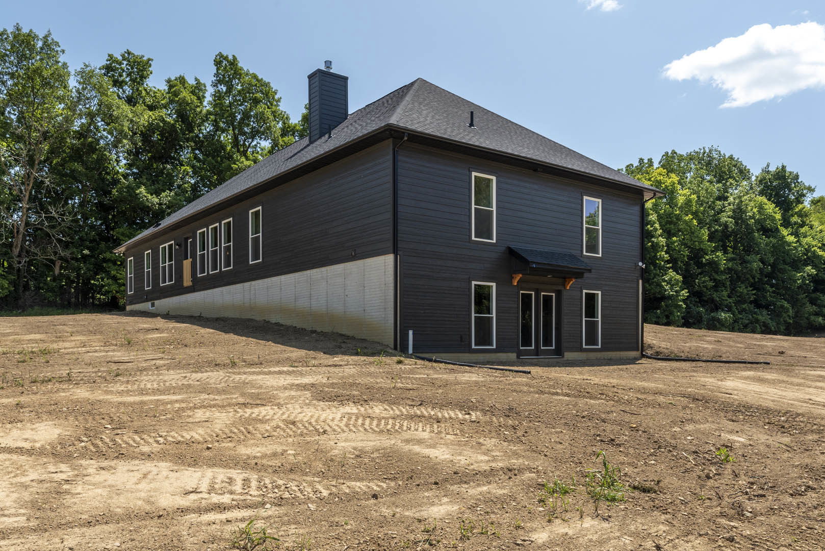 Modern house with black roof, white-framed windows, and light exterior siding, set on a dirt lot with trees and a grassy hill in the background under a partly cloudy sky