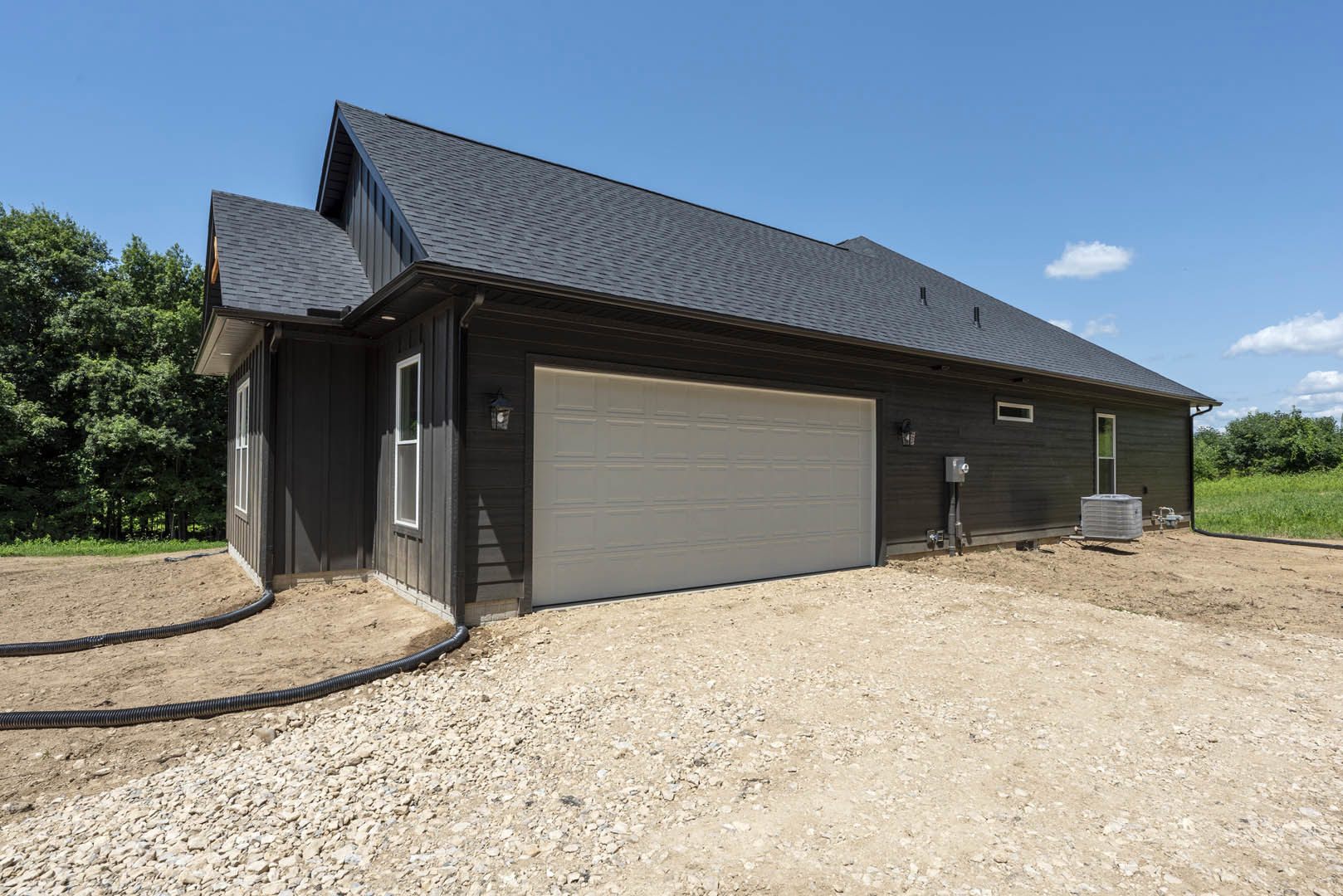 Two-story house with attached garage, white siding, gravel driveway, outdoor lighting, and surrounding trees under a partly cloudy sky