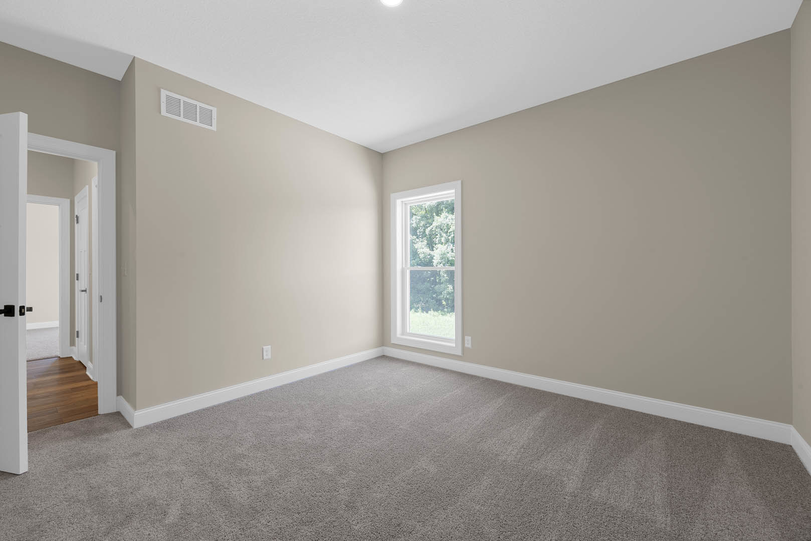 Bedroom with beige carpet, large window overlooking trees, white walls, wood baseboards, and wall vent
