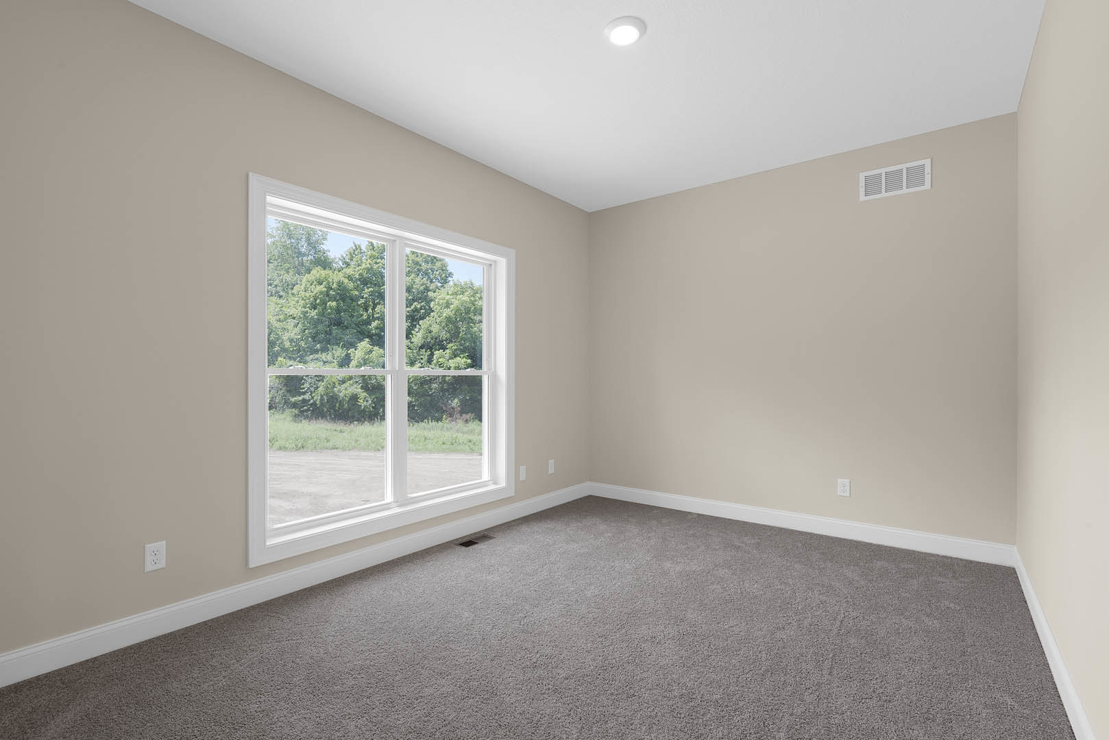 Bright room with white walls, large window overlooking trees, grey carpet flooring, ceiling light fixture, wall vent, and simple baseboard molding