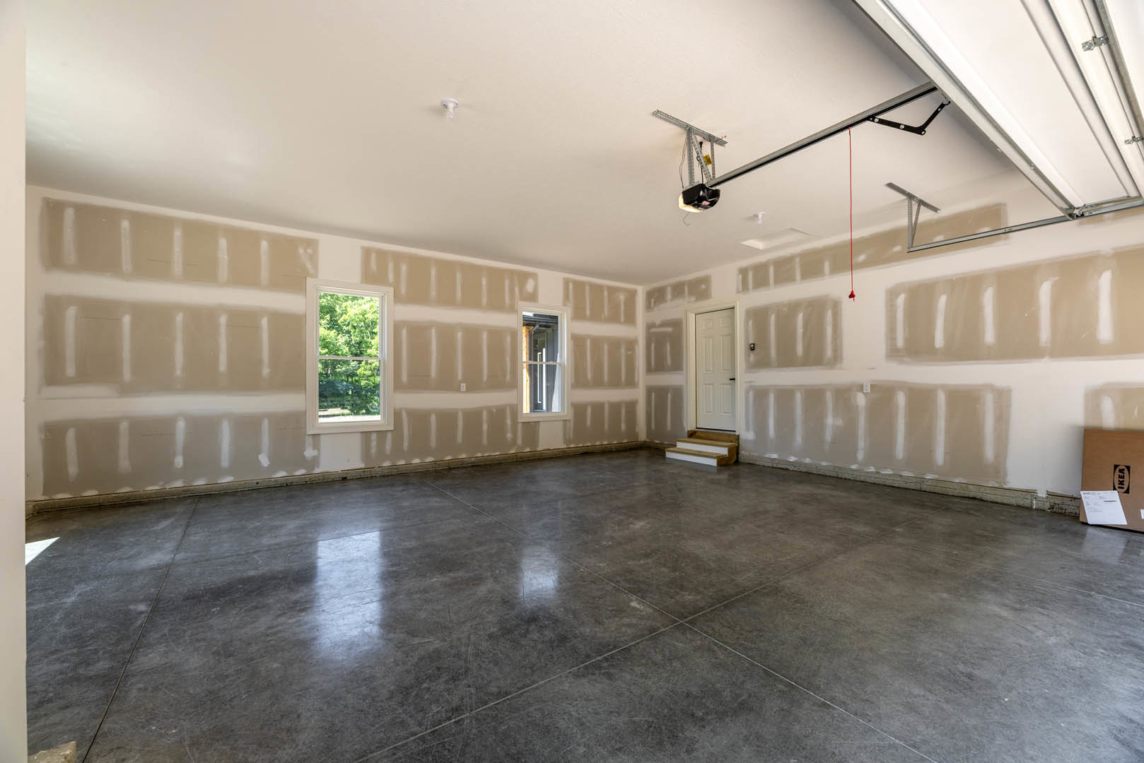 White-walled room with polished concrete floor, large window showing trees outside, white door with black handle, and staircase with grey tile steps