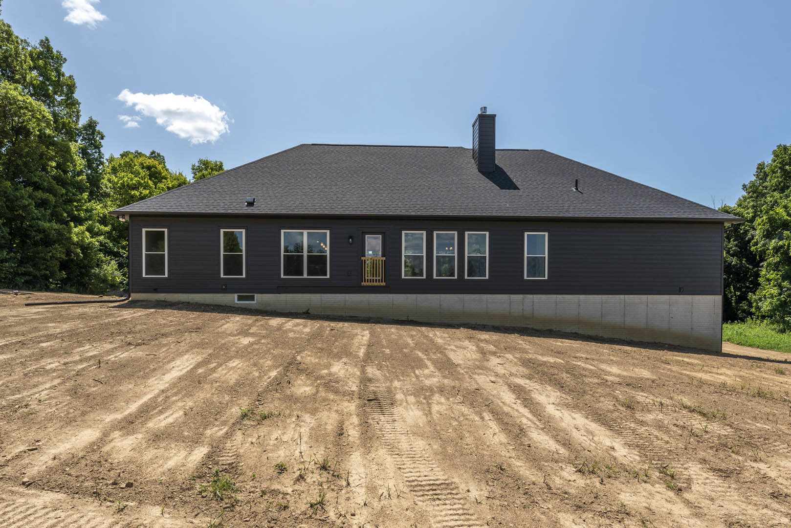 Modern house with black roof sits atop a dirt hill, tire tracks visible in foreground, large windows framed with wood, clouds overhead, cityscape visible through window.