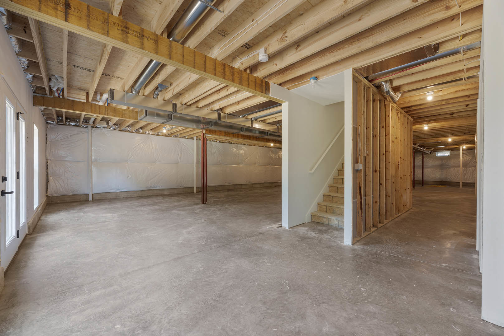 Open room featuring exposed wood beam, concrete floor, staircase with metal railing, white wall partially covered by plastic sheeting, and close-up of modern door handle