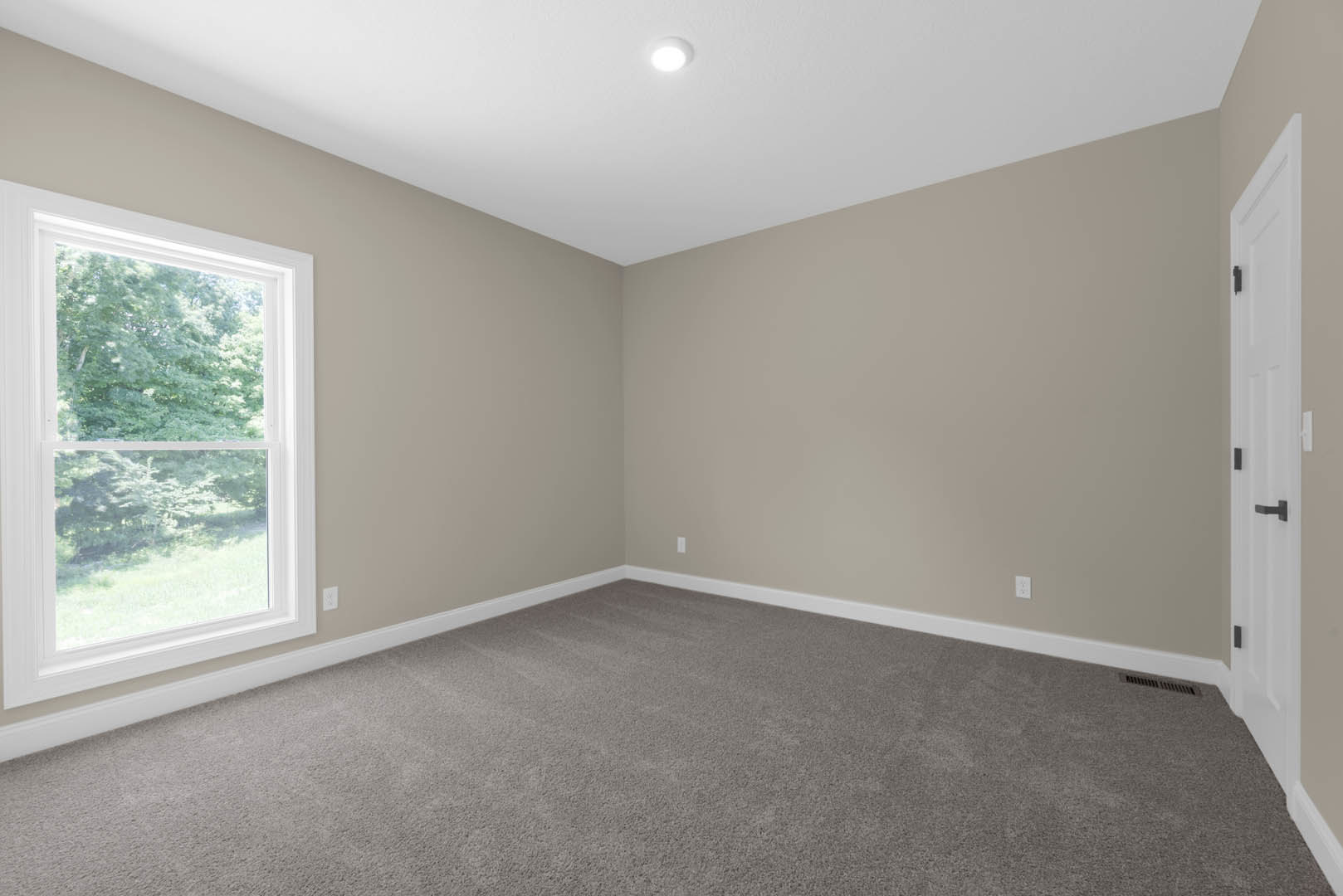 Bright room with large window overlooking trees, grey carpet flooring, white walls with molding, recessed ceiling light, and white door featuring a black handle