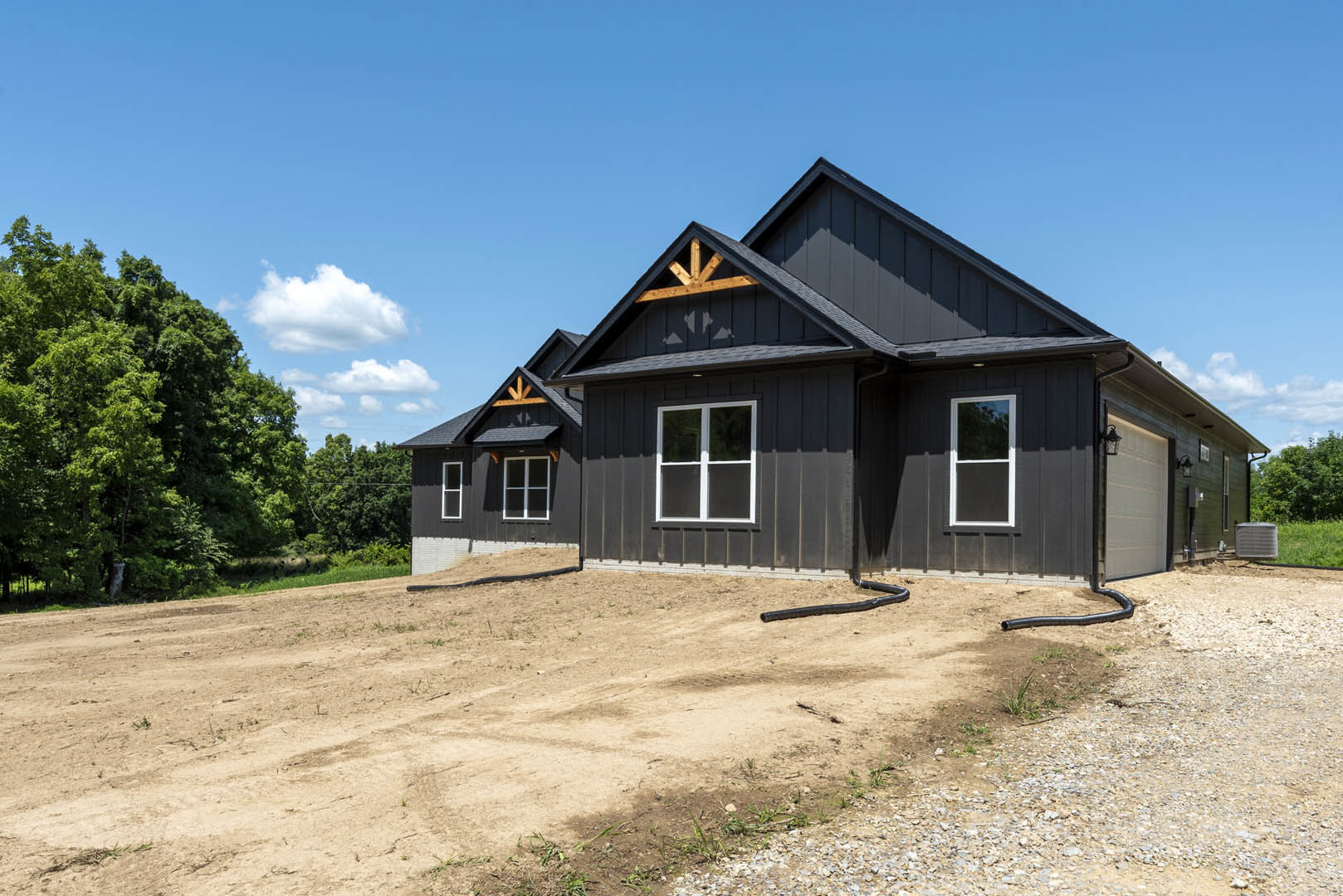 Two-story house with gray siding, white trim windows, shingled roof, central air conditioning unit, surrounded by bare dirt lot and mature trees in the background