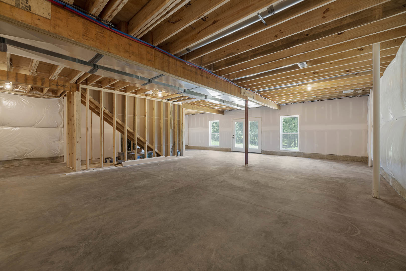 Open room with exposed wood beam ceiling, concrete floor, central metal support pole, white mattress near double glass doors, large window showing trees outside