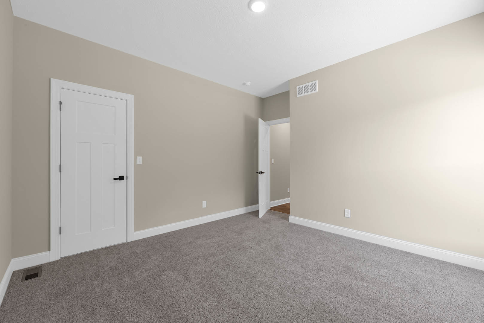 Beige-walled room with white door featuring black handle, carpeted floor, ceiling and wall vents, simple molding