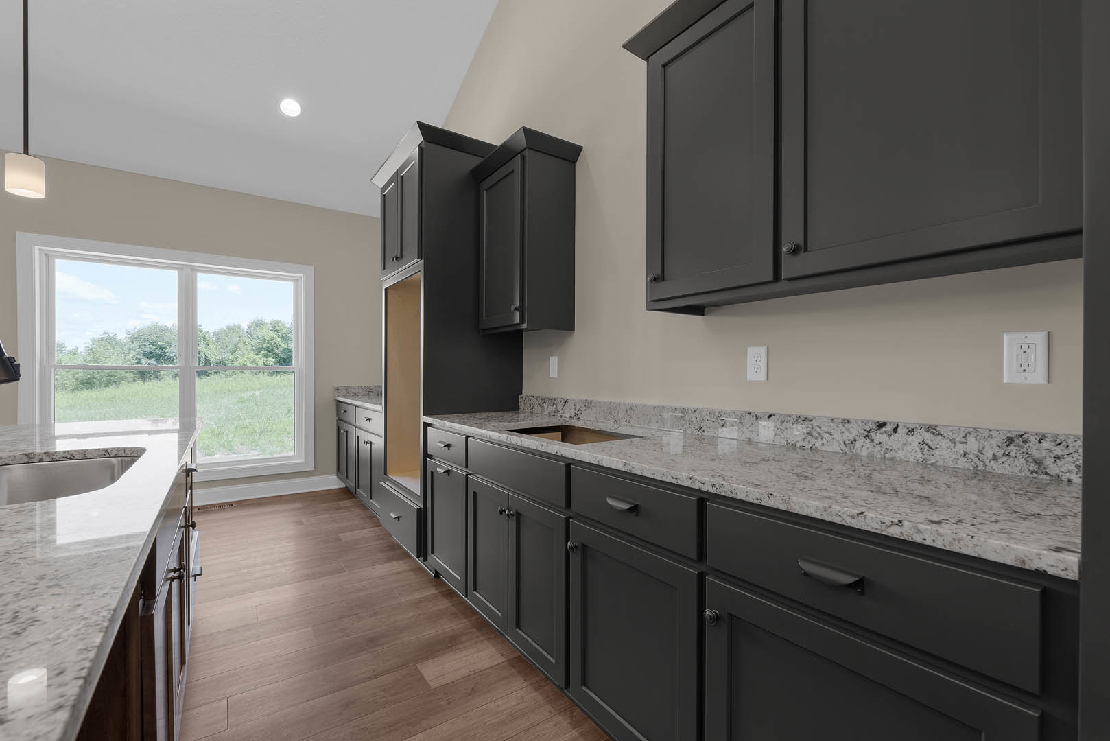Black kitchen cabinets with brushed metal handles, light wood flooring, white walls, stainless steel sink, and a large window overlooking the outdoors.