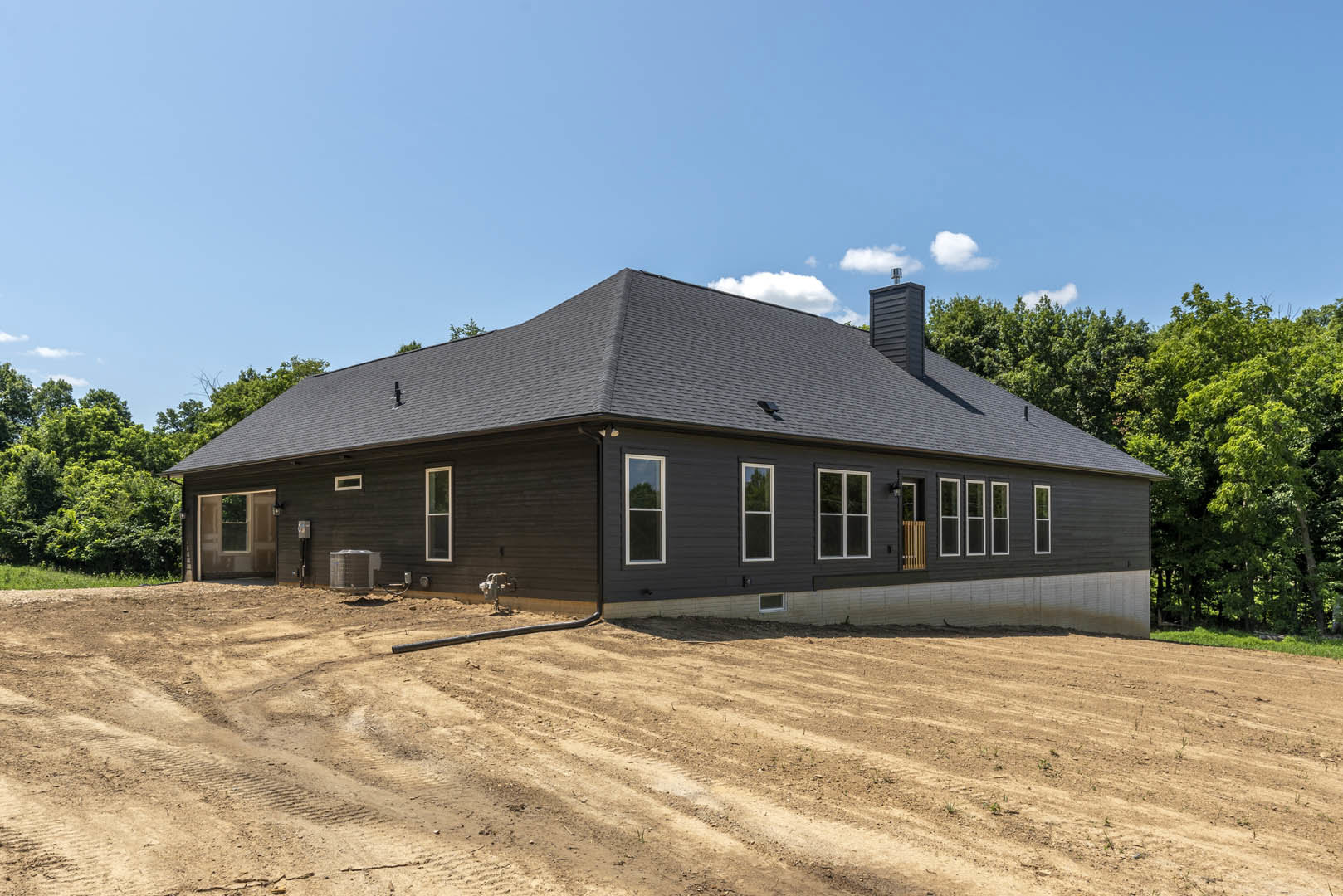 Partially built house with black roof, exposed framing, large metal container on dirt lot, scattered construction materials, trees and blue sky in background