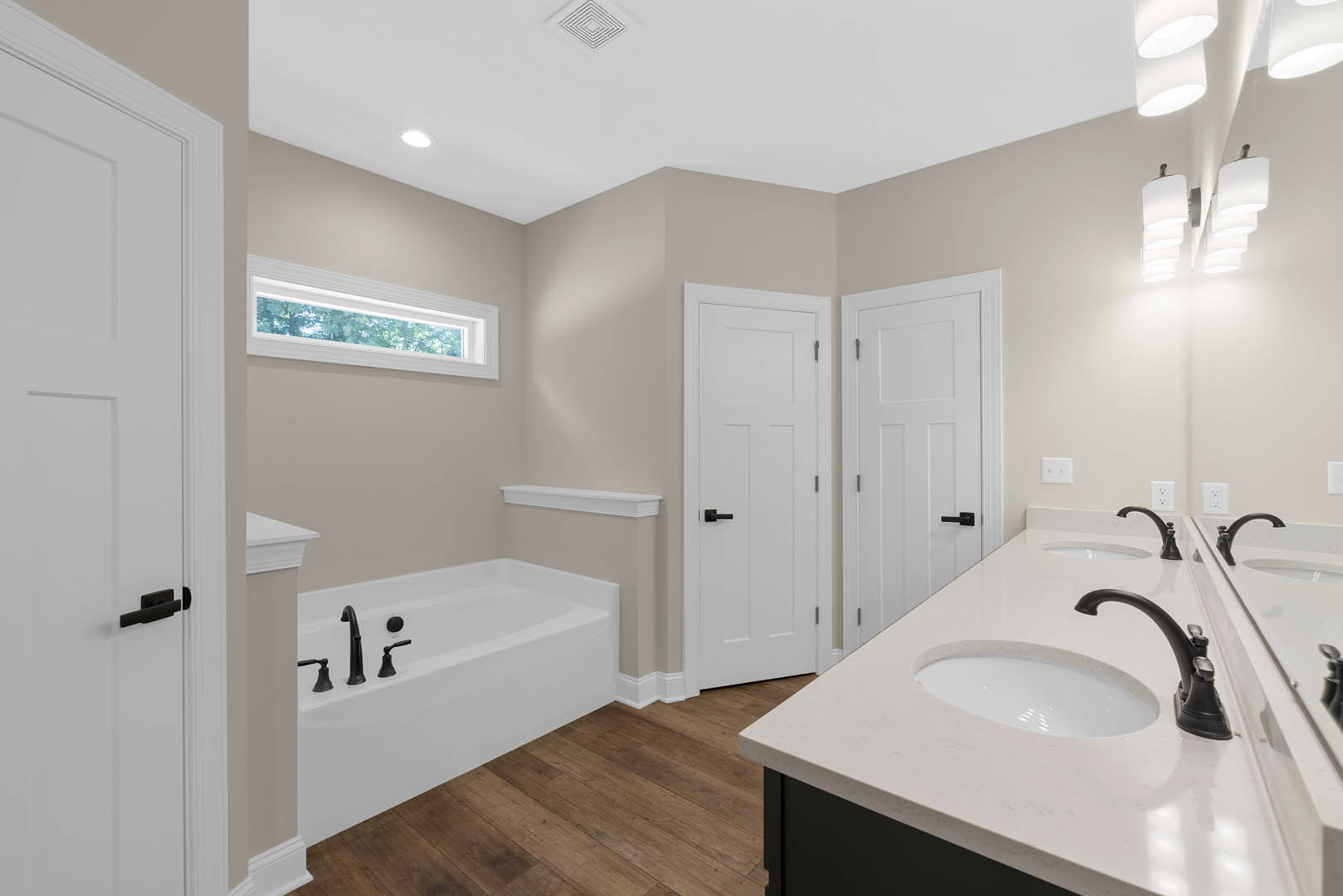 Bathroom with freestanding bathtub, dual sinks with chrome faucets, wood flooring, white door with black handle, and rectangular window letting in natural light