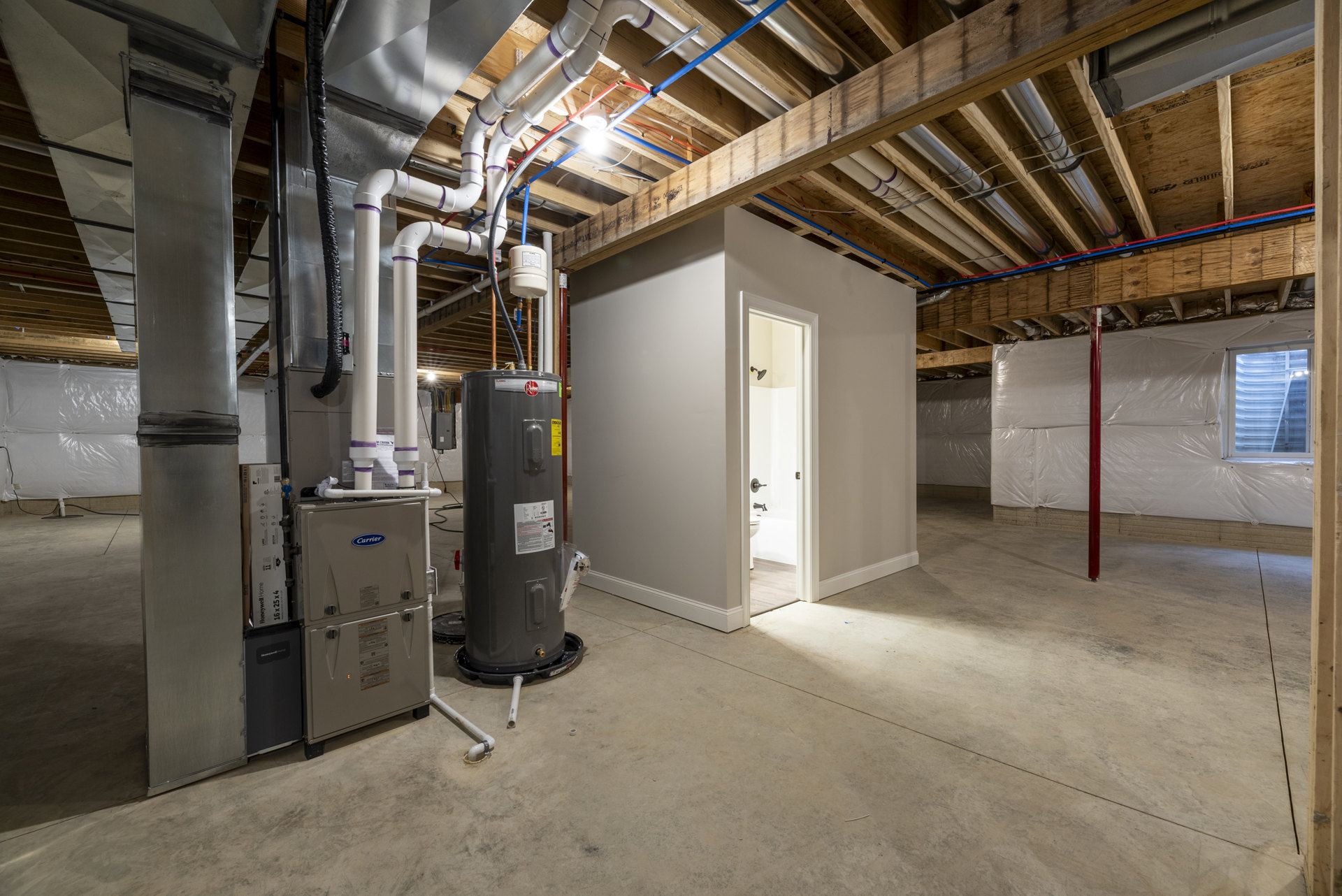 Unfinished basement with exposed steel beams, visible pipes along ceiling and walls, water heater, white-framed window, open bathroom door, and white container with blue lid on